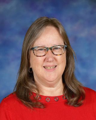 A woman wearing glasses and a blue sweater is standing in front of a tree.