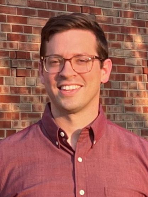 A man wearing glasses and a red shirt is standing in front of a brick wall.