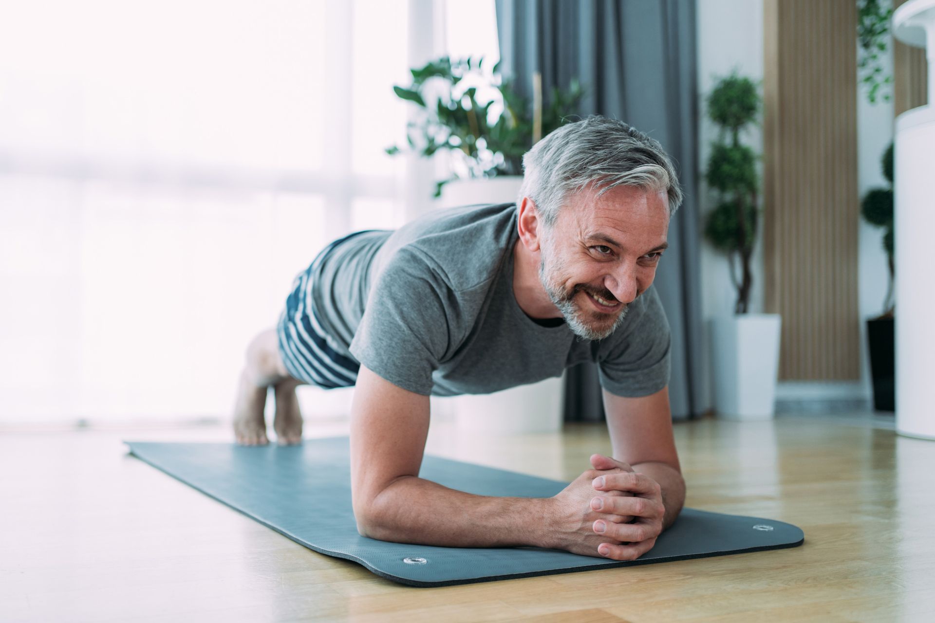 Man on an exercise mat, performing a plank in a sunlit room, smiling.