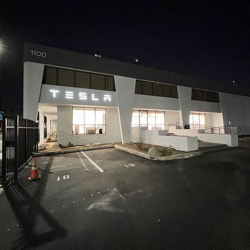 A tesla building is lit up at night with a parking lot in front of it.