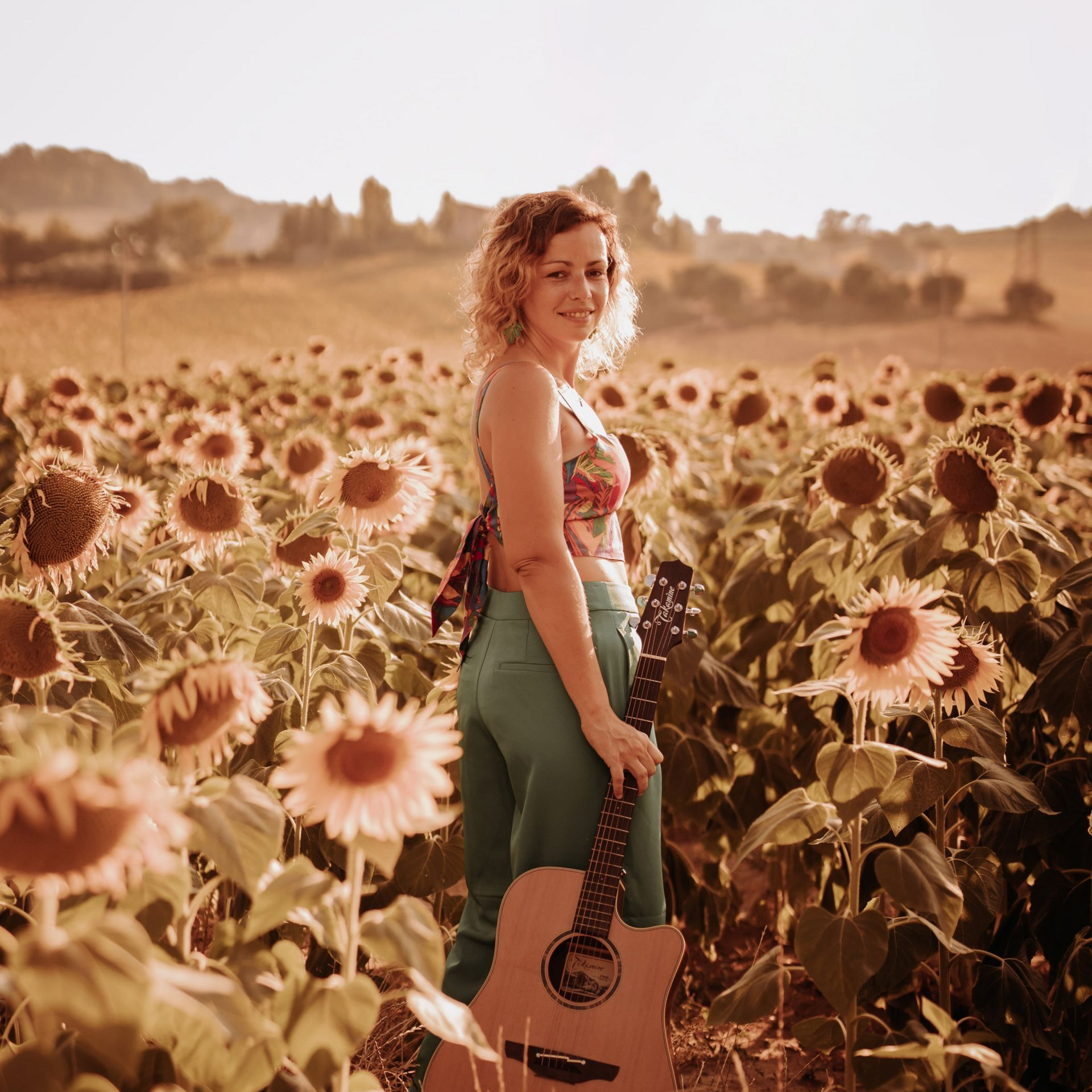 Woman with guitar in a sunflower field, wearing green pants and a floral top, smiling.