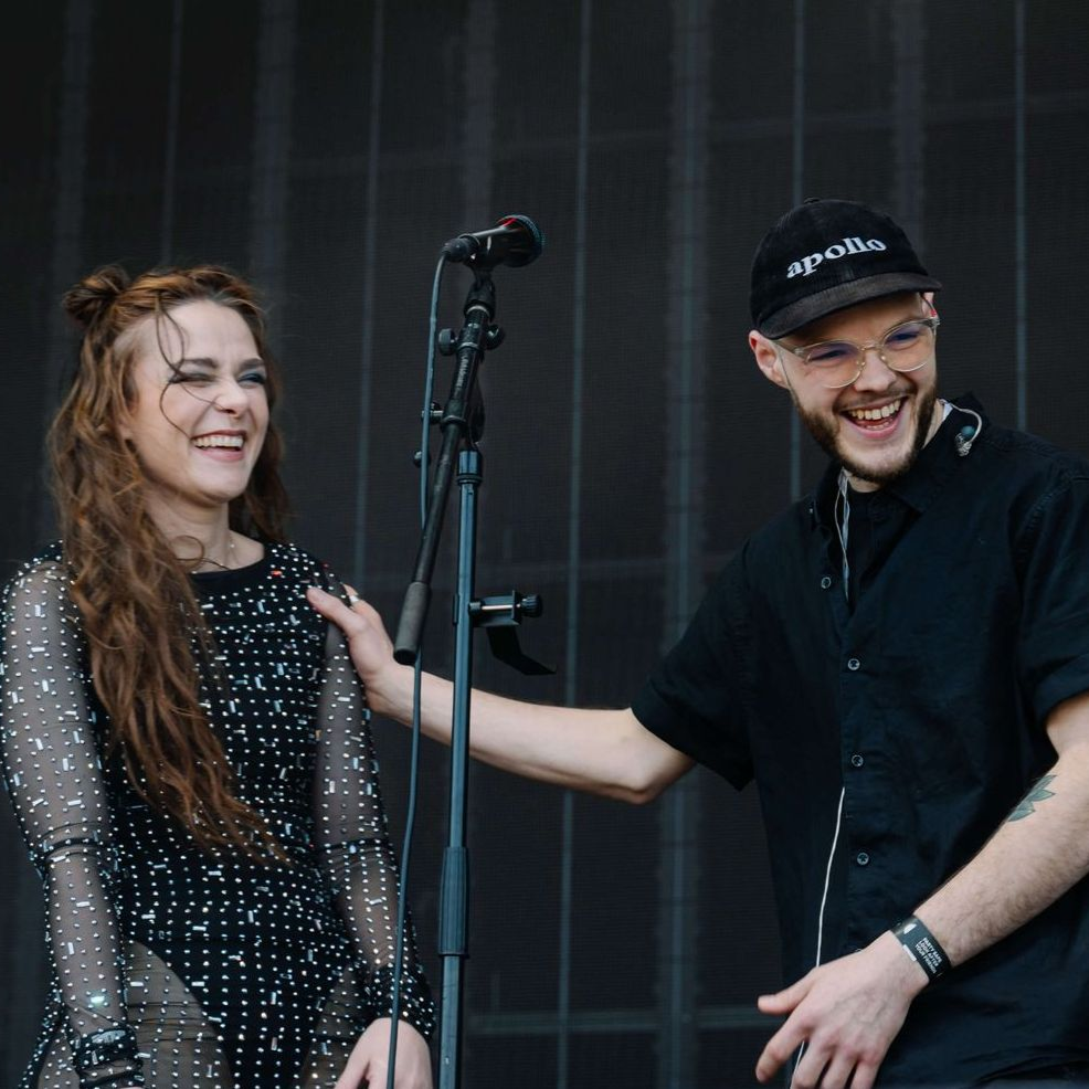 A woman in black outfit and a man in a black shirt stand laughing next to a microphone on stage.