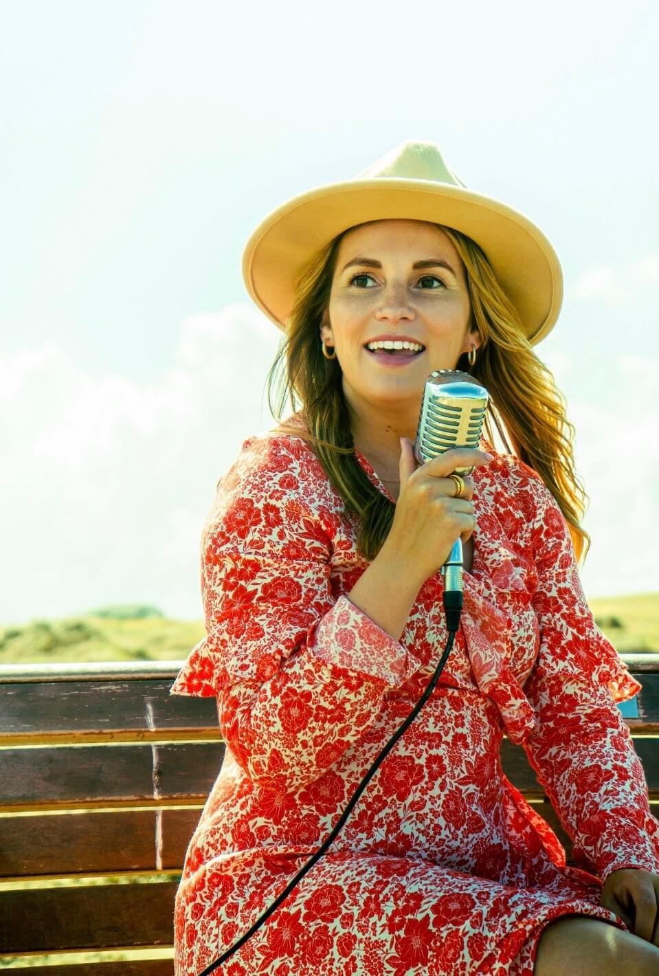 A woman in a hat is holding a microphone while sitting on a bench.