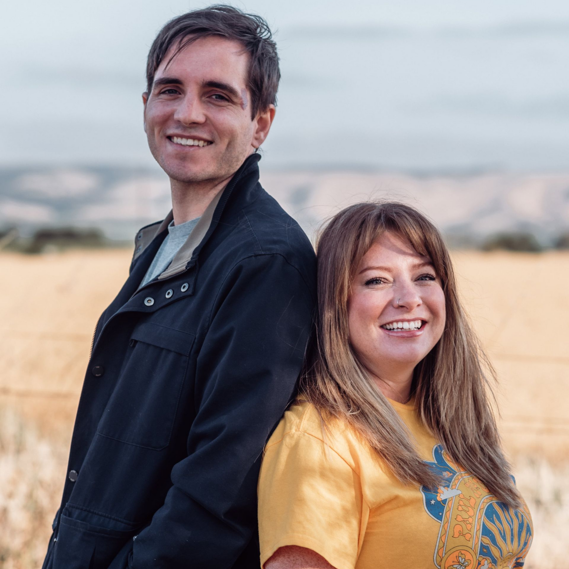 Man in black jacket and woman in yellow shirt smile, standing back to back outdoors.