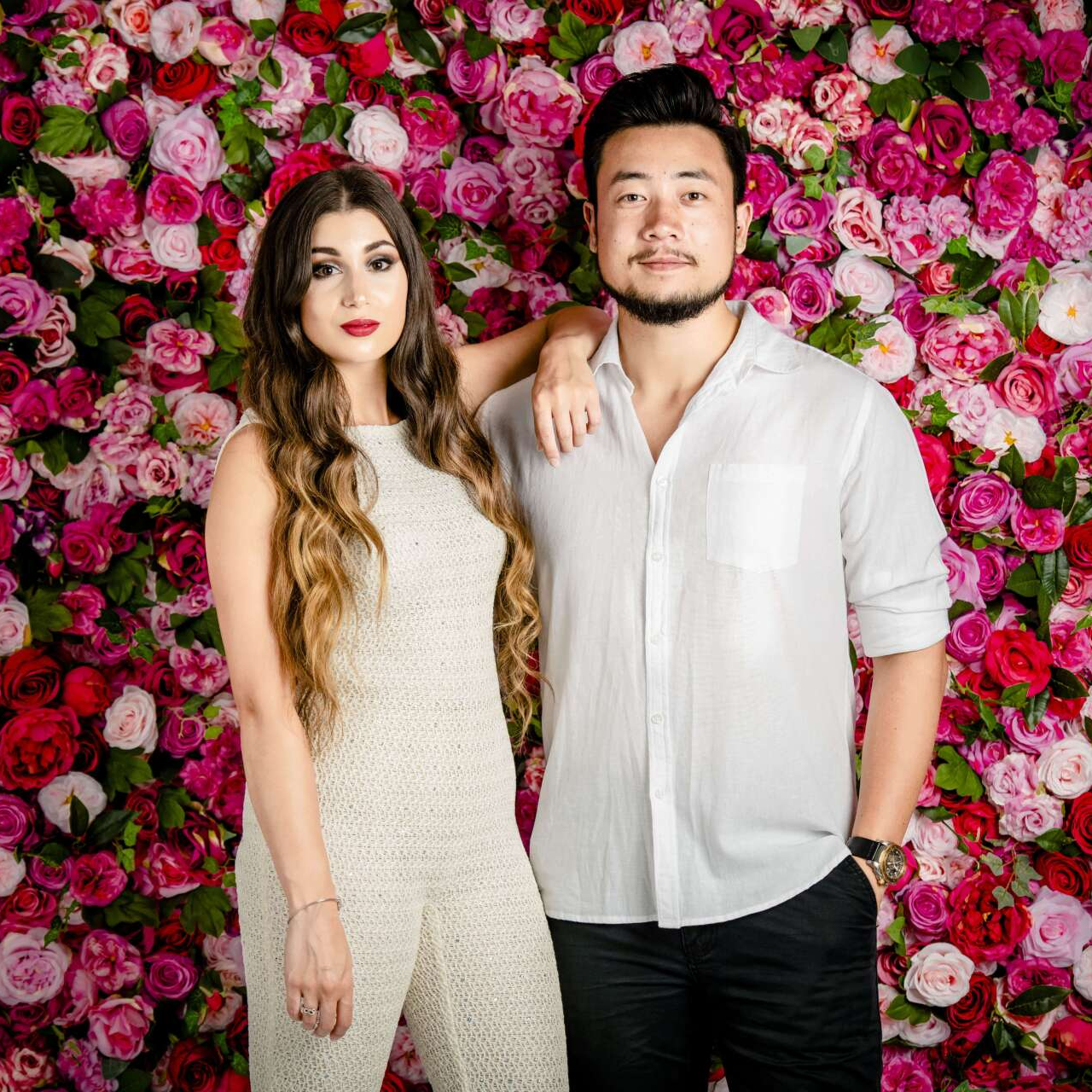 Woman in jumpsuit and man in button-down shirt pose in front of a floral backdrop.