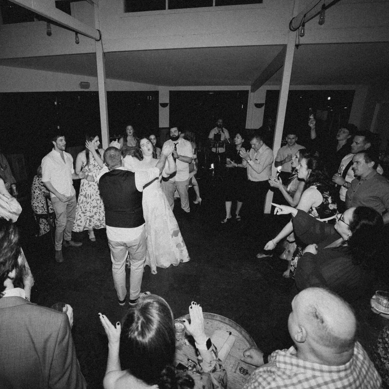 Black and white photo of a wedding reception. Bride and groom dance, surrounded by guests taking photos and watching.
