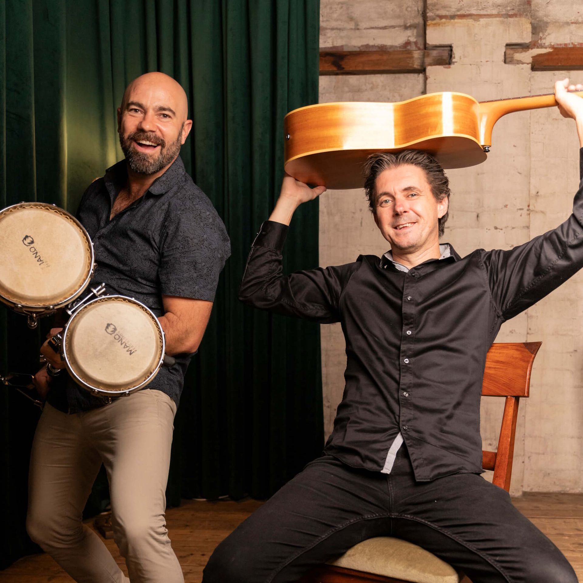 Two men smiling, one holding bongos, the other holding a guitar over his head.