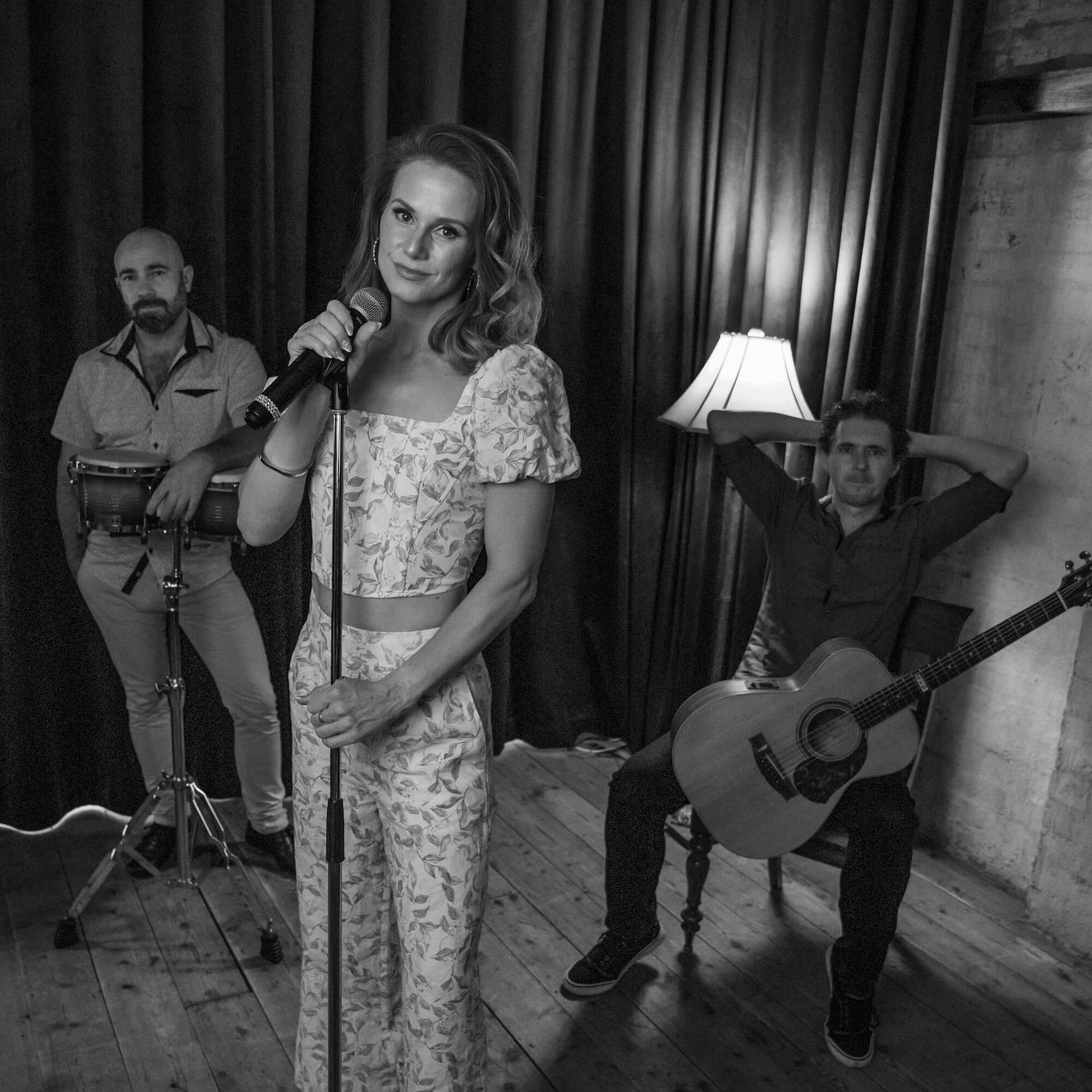 Band performing: Woman singing, man playing drums, guitarist. Black and white, casual setting.