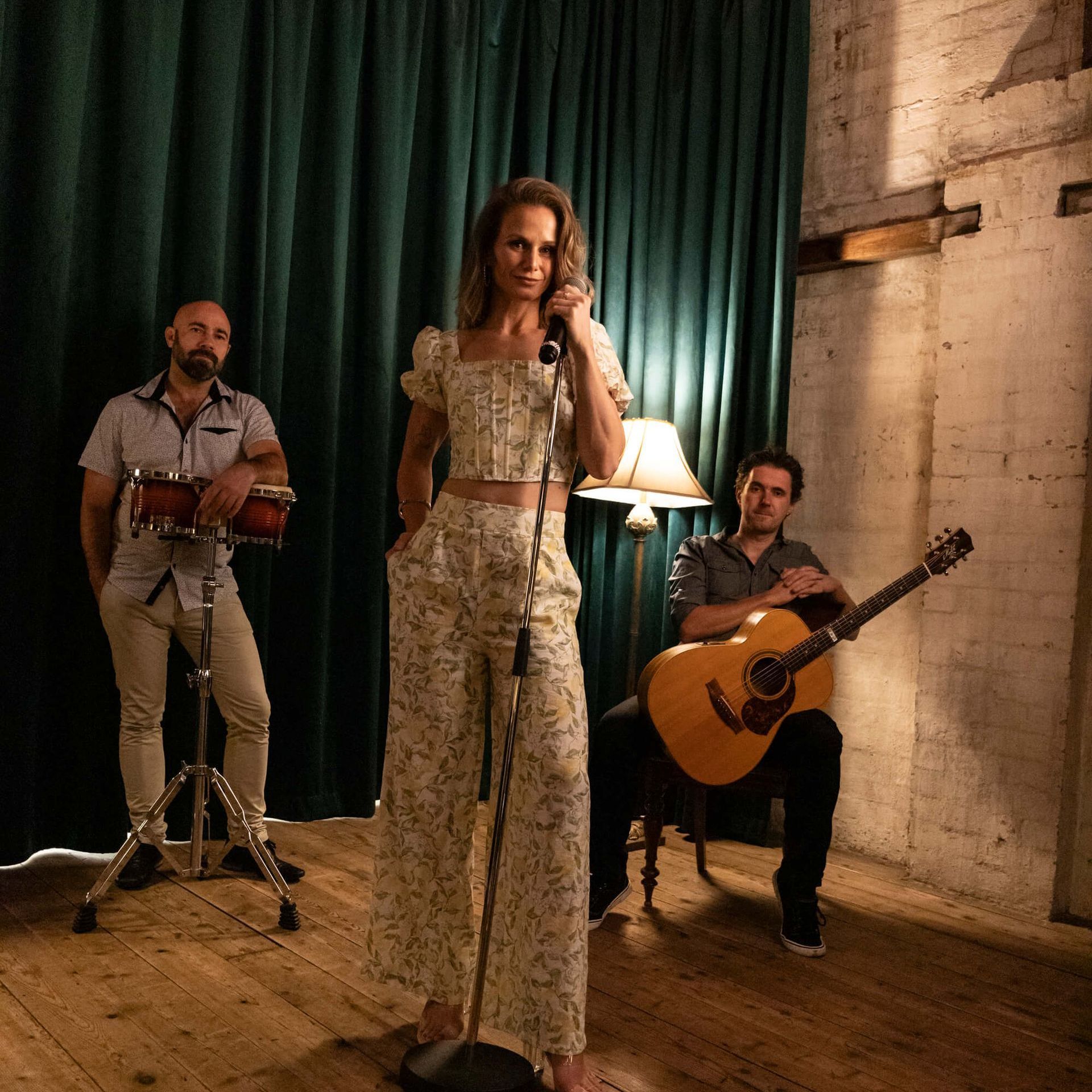 A musical trio performing: woman singing, guitarist, conga player. In a room with curtain and exposed brick.