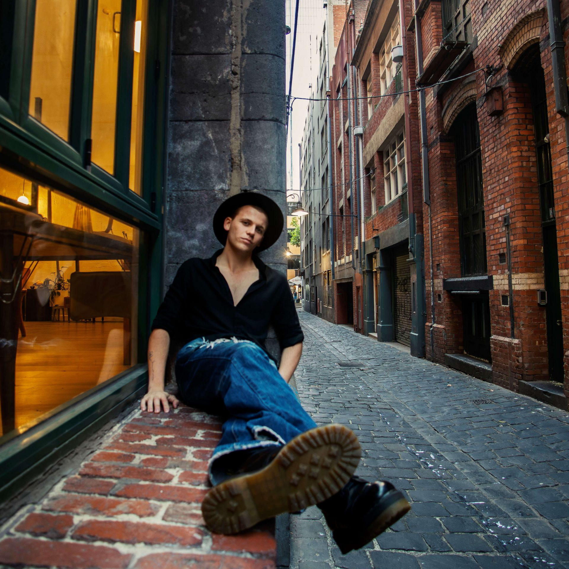 Person sitting on a cobblestone alley ledge beside brick buildings, wearing a black hat and blue jeans.
