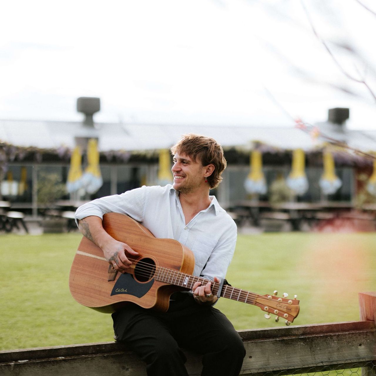 Man with guitar, smiling, sitting on fence outdoors. Green grass and building in background.