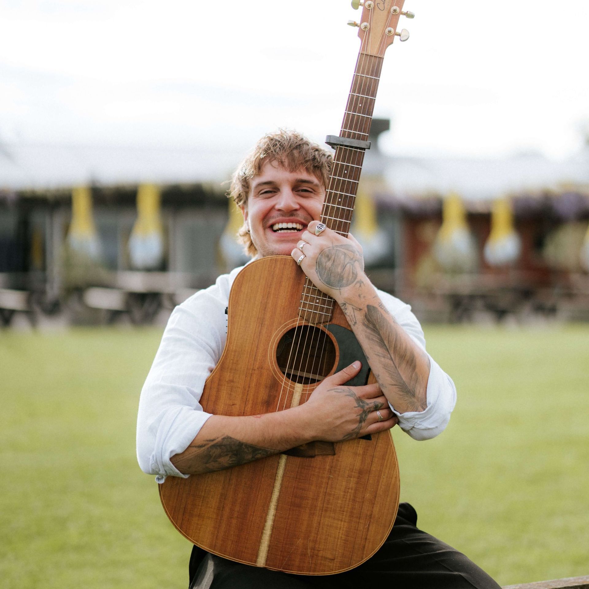 Man with tattoos smiles, holds acoustic guitar outdoors in front of blurred building.
