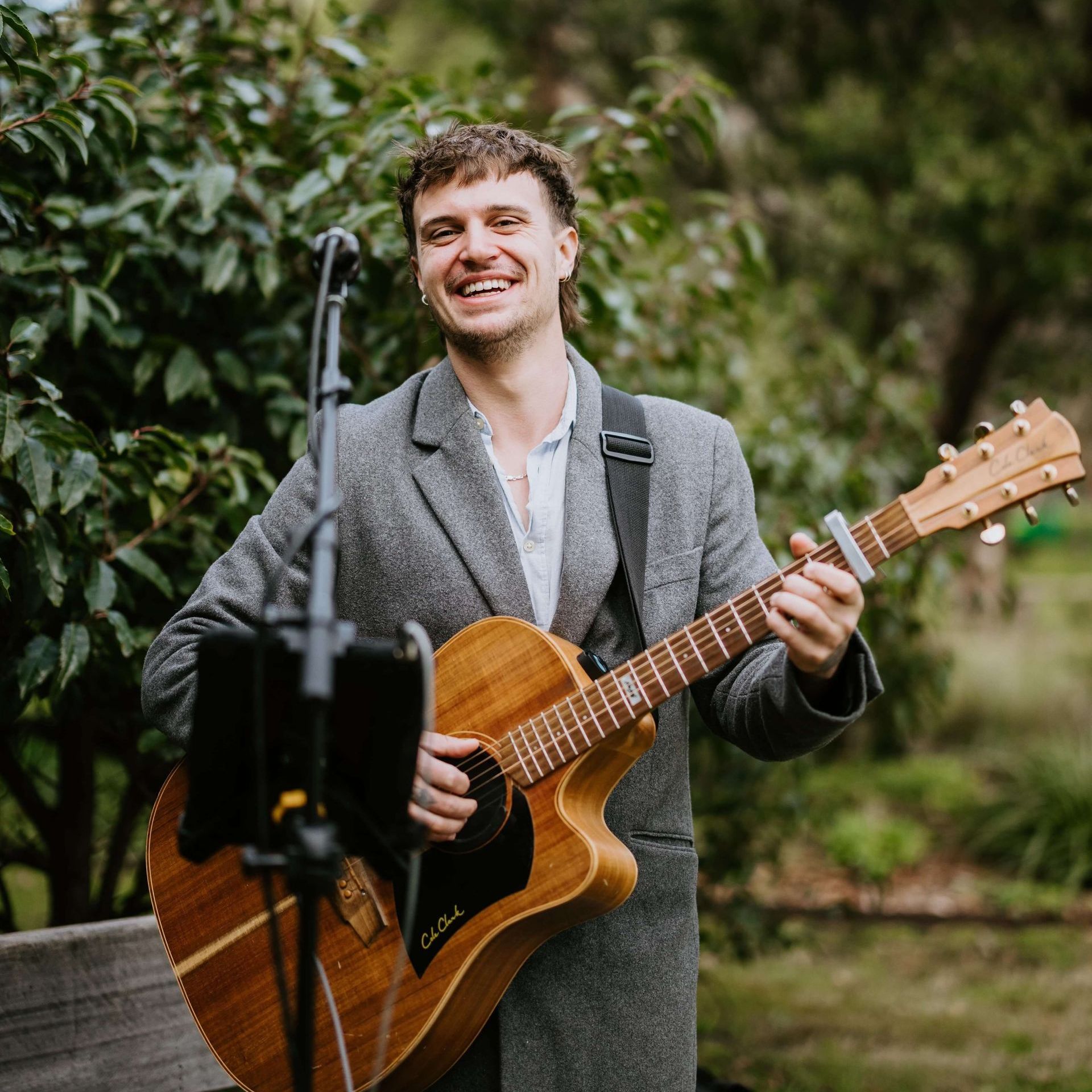 Man with guitar, smiling, performing outdoors in a gray coat.