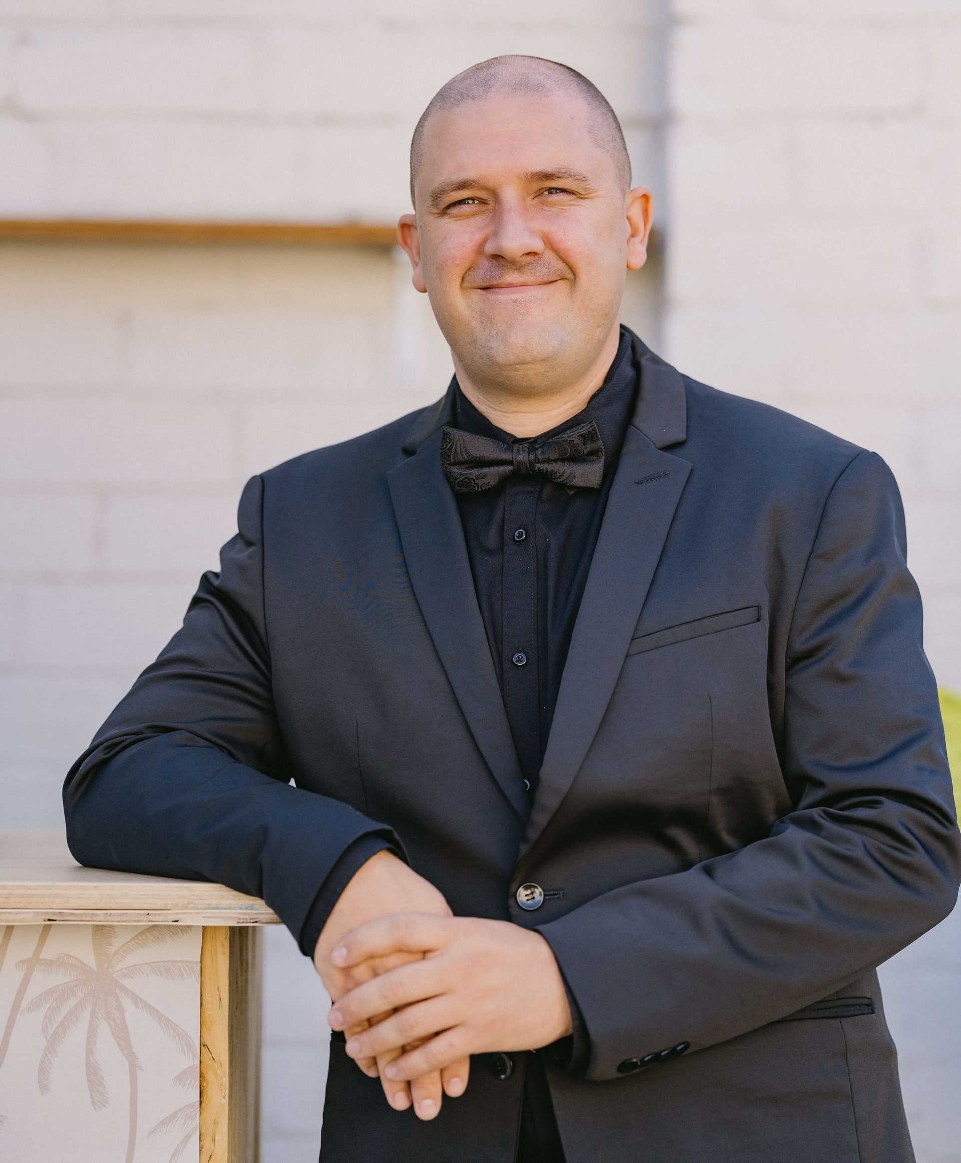 Man with bald head, in black suit and bow tie, leaning on a wooden structure, smiling outside.