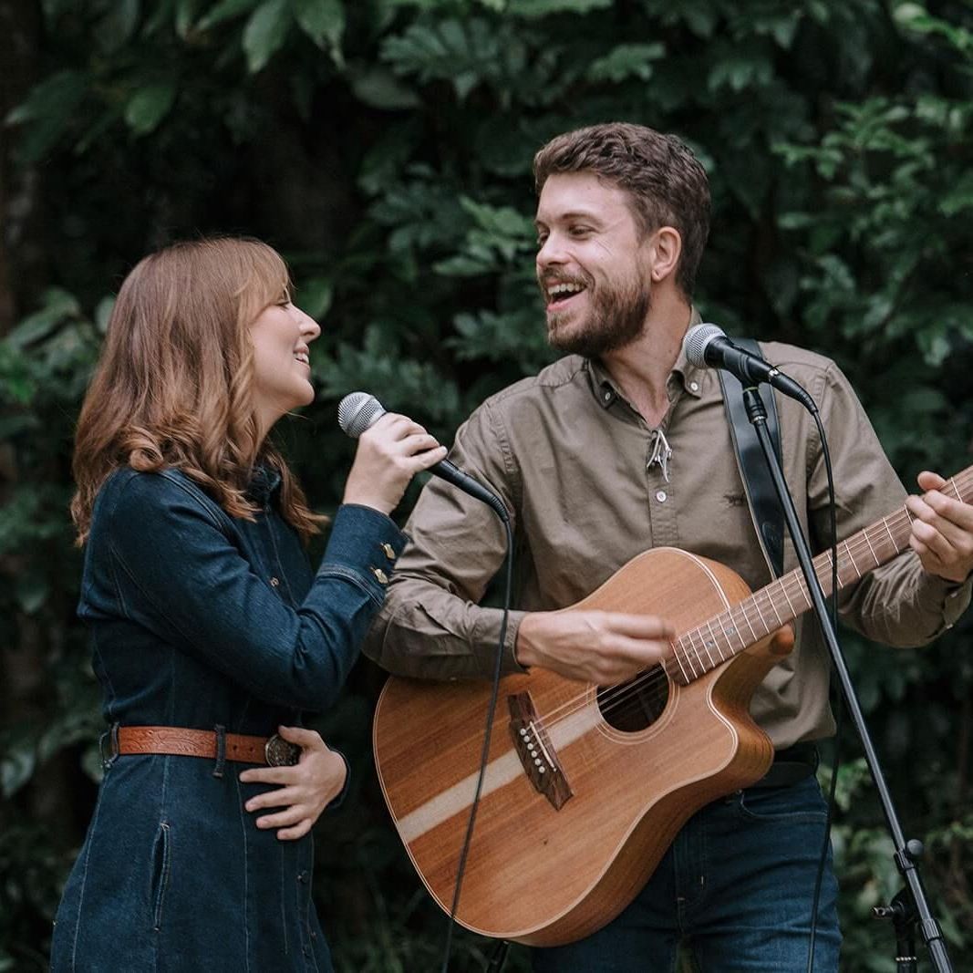 A woman and a man sing and play acoustic guitar outdoors; green foliage in the background.