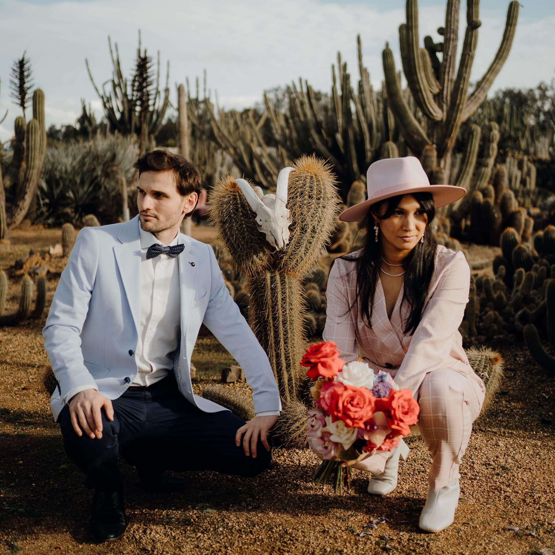 Couple in formal wear kneels by cactus in desert garden; woman holds flowers, man looks left.