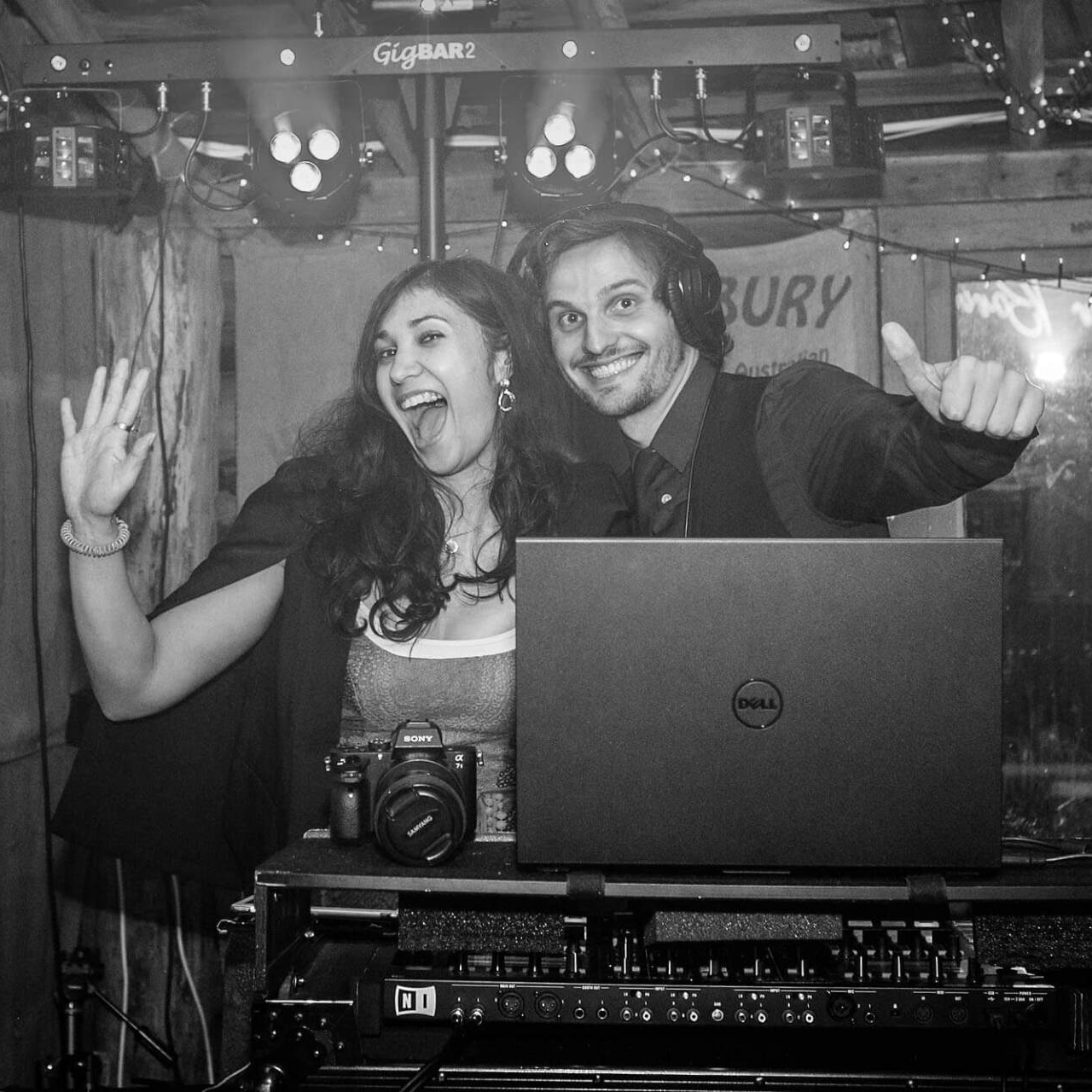 Two DJs at a party. Woman waves, man gives thumbs up.  Black and white, DJ equipment present.
