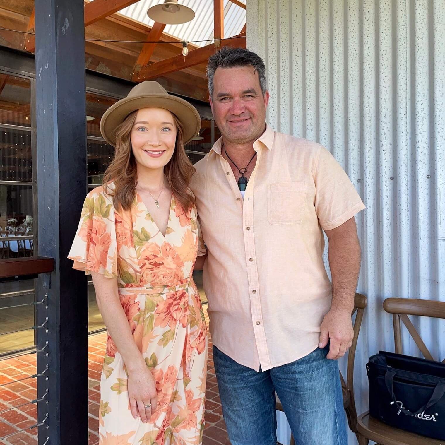 Woman in floral dress and hat, man in peach shirt and jeans, smiling, posing outside.