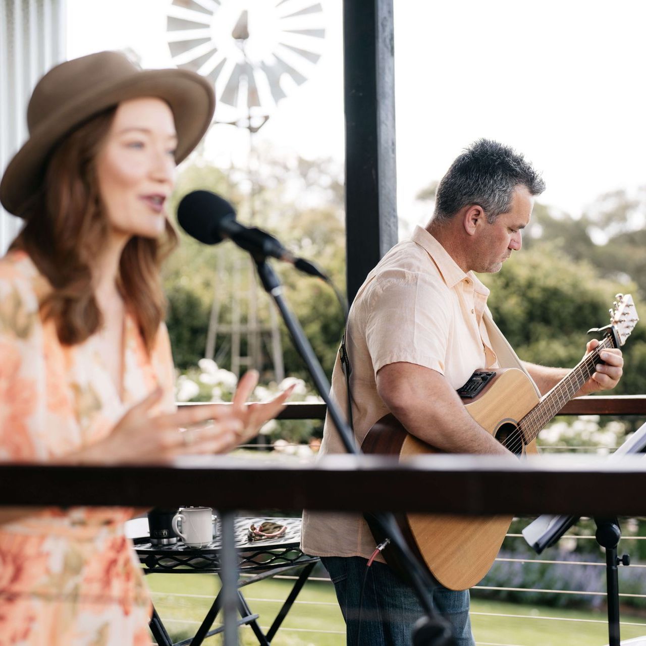 Woman singing into a microphone and man playing guitar on a porch, both dressed casually, with a windmill in the background.