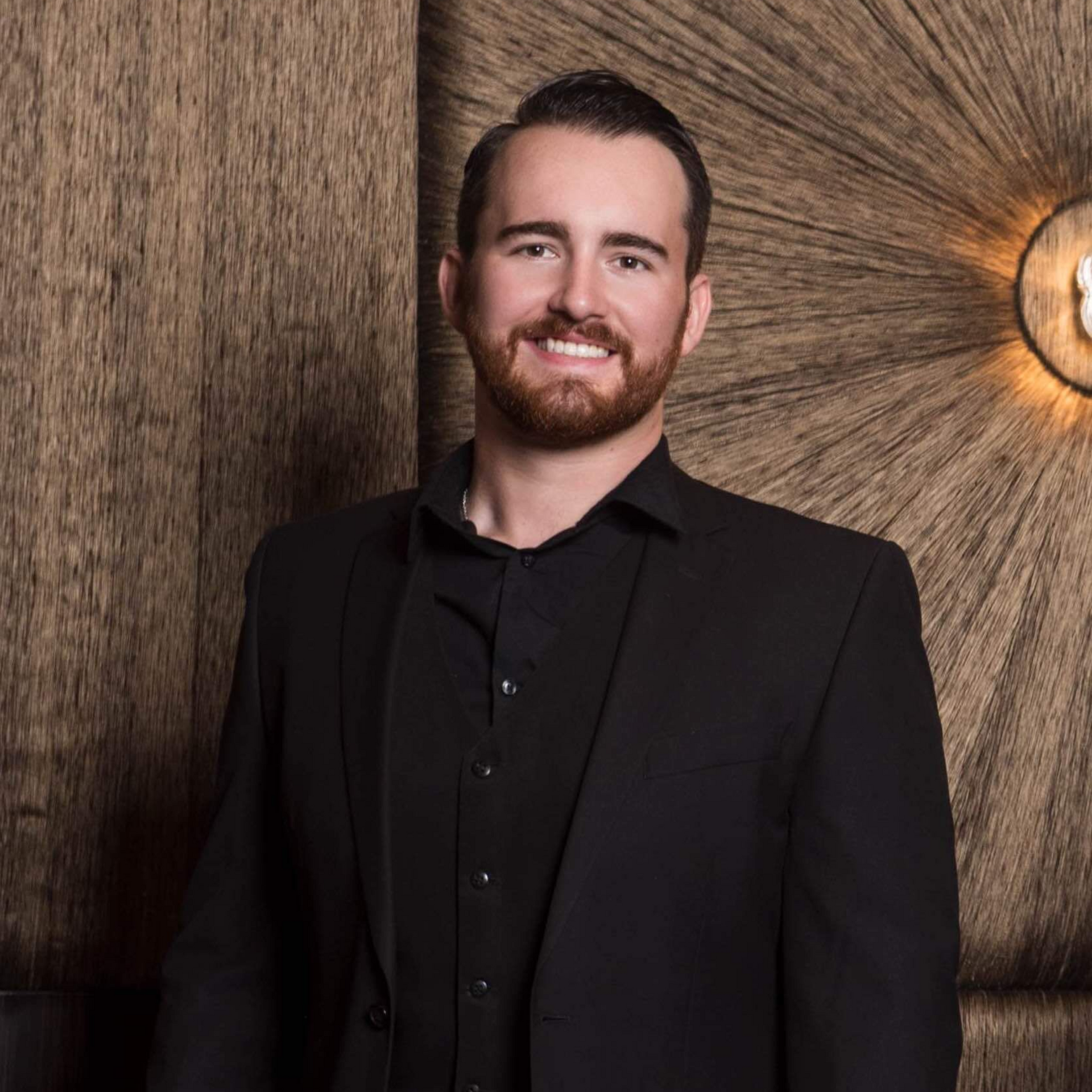 Man in black suit smiles, standing near textured wooden wall.