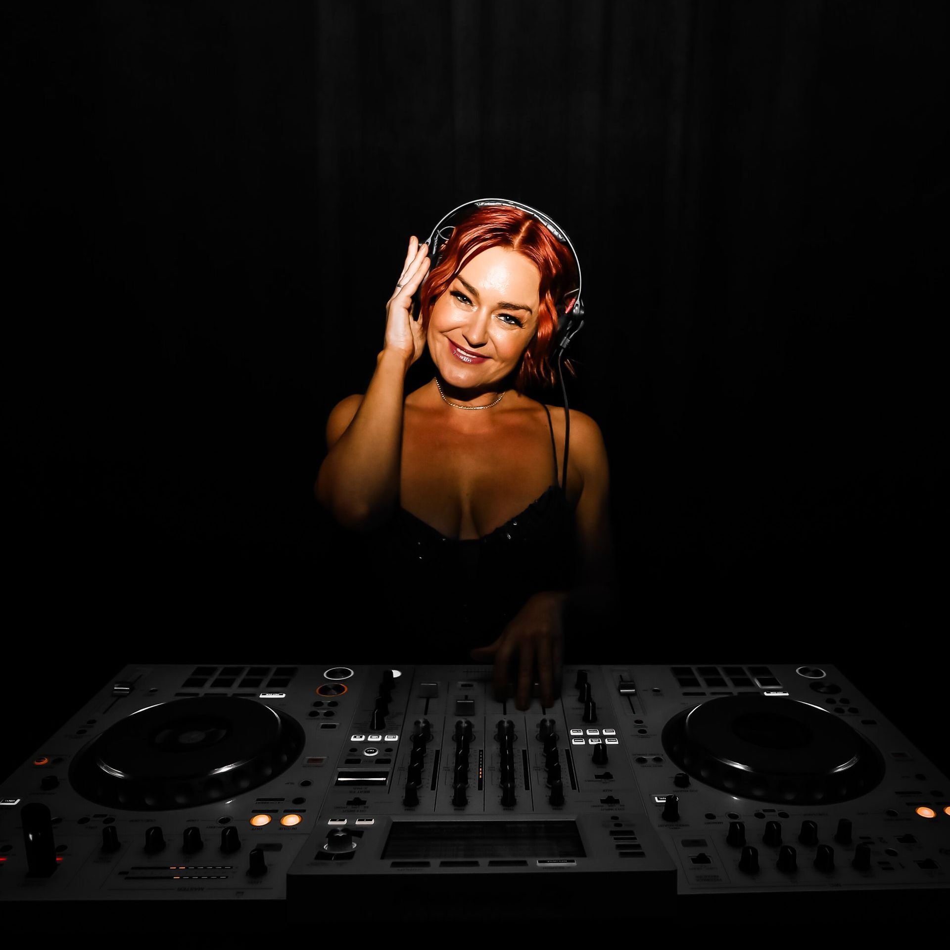 Woman DJ with red hair, smiling, wearing headphones, at a console in a dark setting.