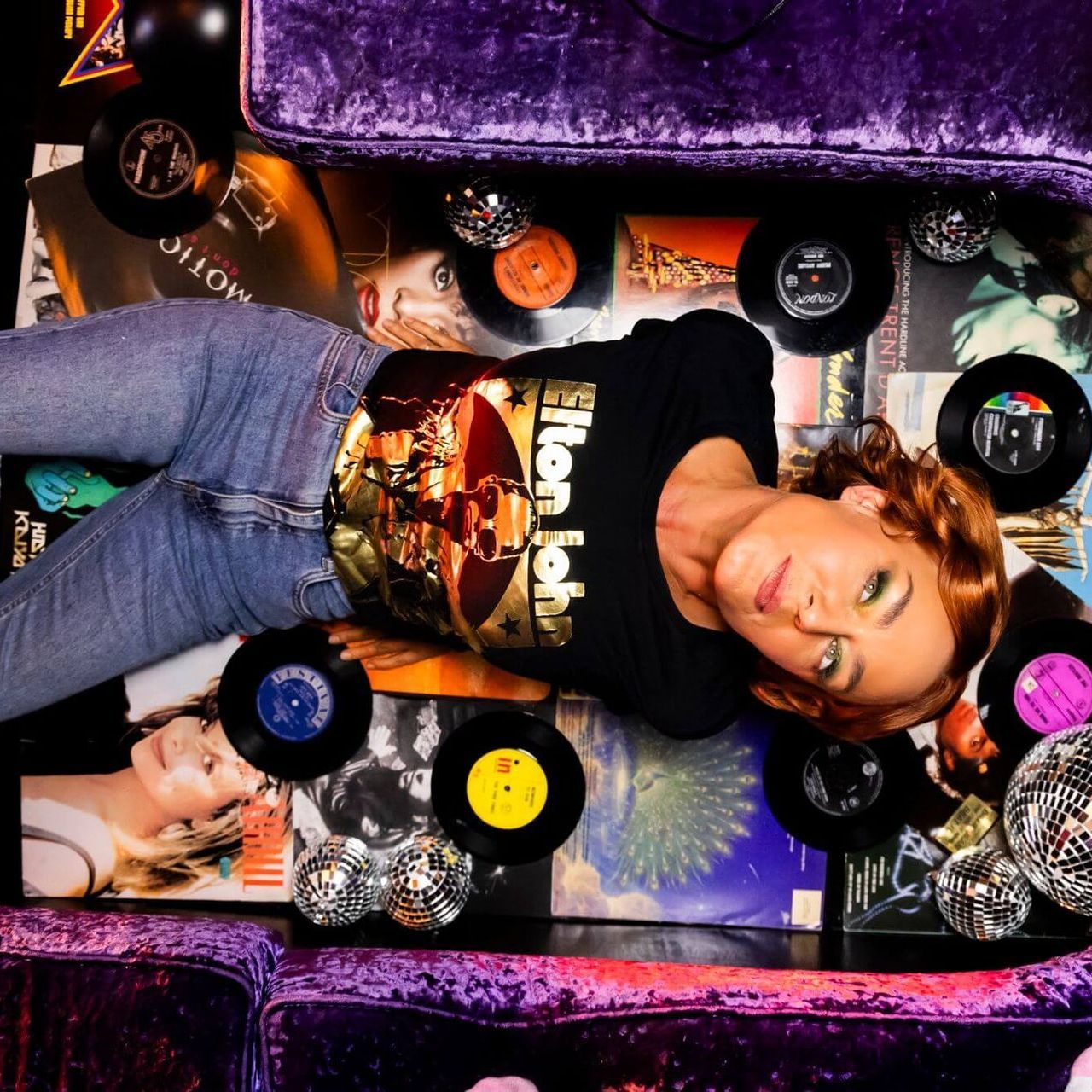 Woman in Elton John shirt on a purple velvet couch surrounded by records and disco balls.