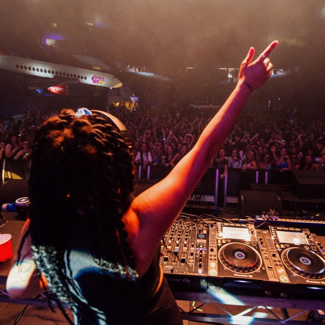 DJ at a concert, arm raised towards crowd, mixing on a console. Airplane is in the background.
