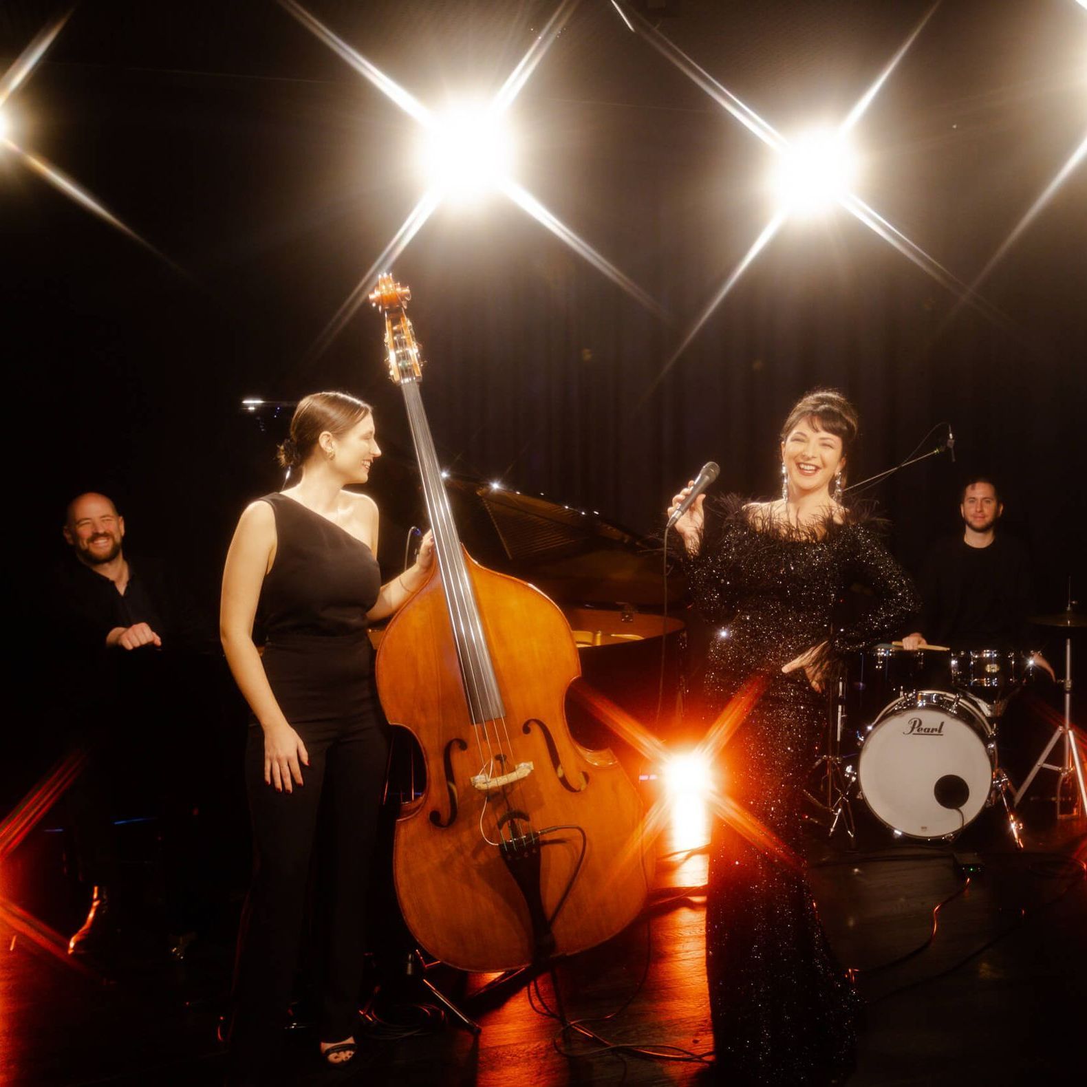 Jazz band performing on stage; woman singing with microphone, upright bass, drums, and piano.