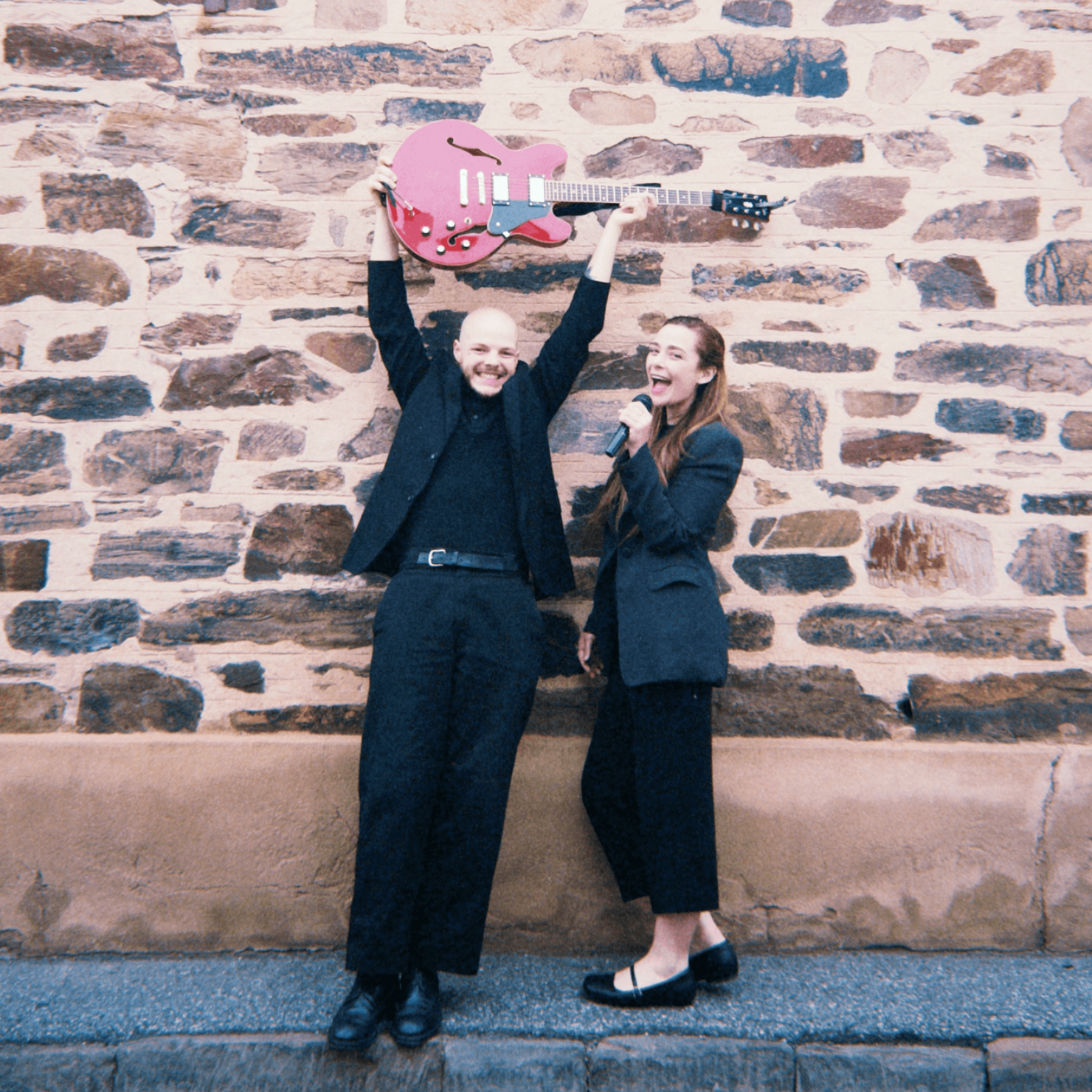 Two people in dark clothes smiling, one holding a pink guitar overhead against a stone wall.