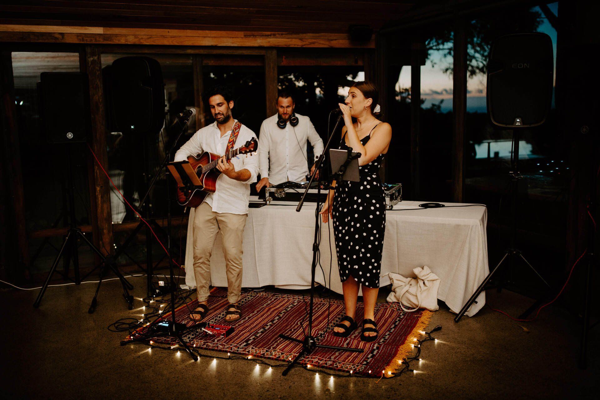 Band performing on a rug at an outdoor event. A woman sings, a man plays guitar, and another DJ’s.