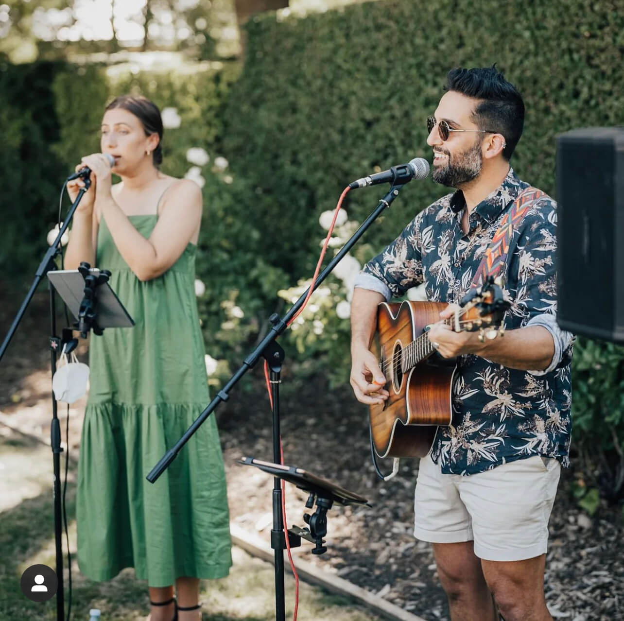 Two musicians perform outdoors: woman in green dress sings, man plays guitar in shorts and floral shirt.