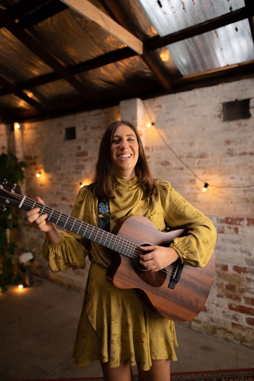 Woman in yellow dress plays acoustic guitar, smiling in a brick-walled room with string lights.