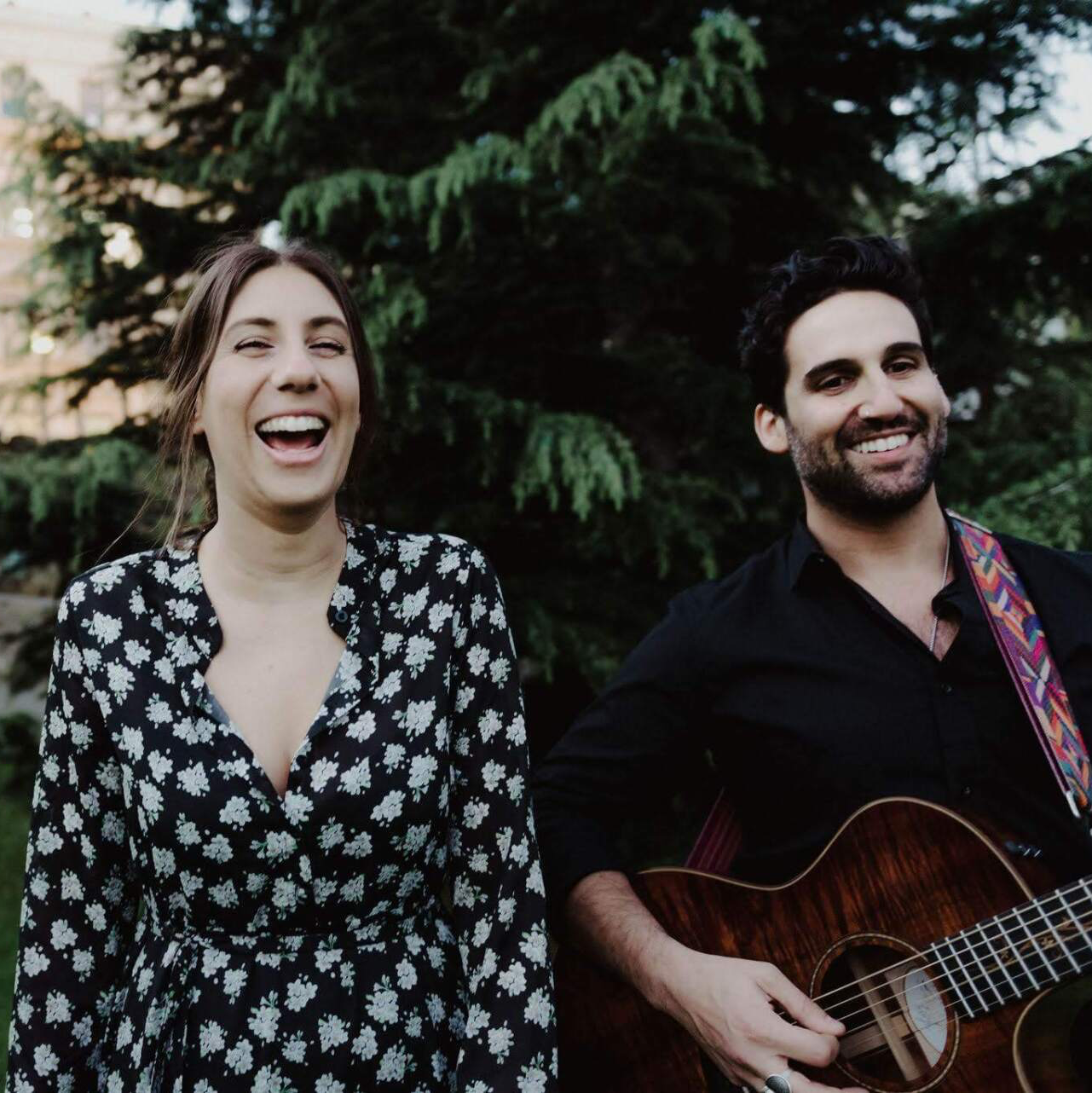 Woman laughing, man playing guitar outside.  Both smiling. Green foliage in the background.