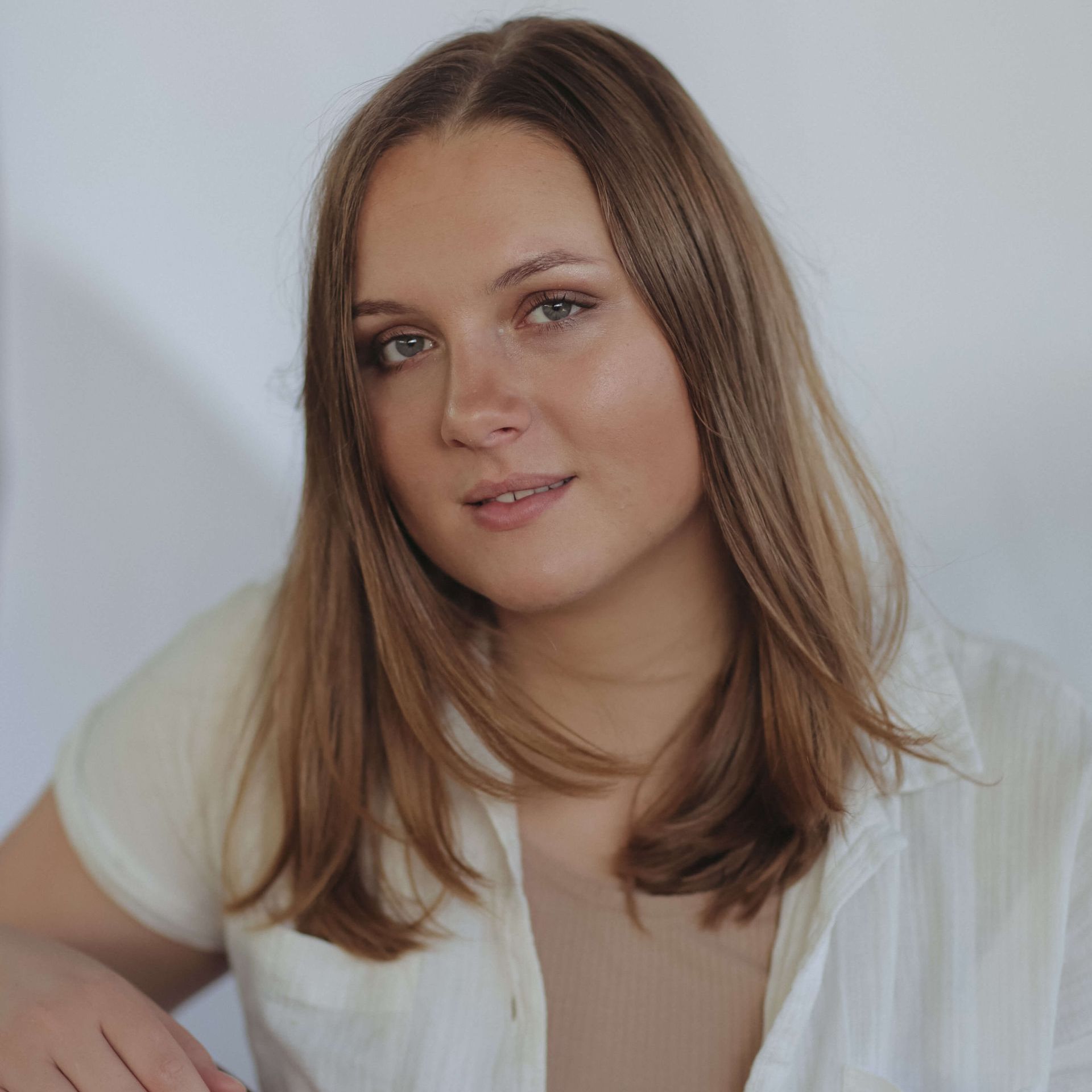 Woman with long light brown hair in a white shirt, smiling against a white backdrop.