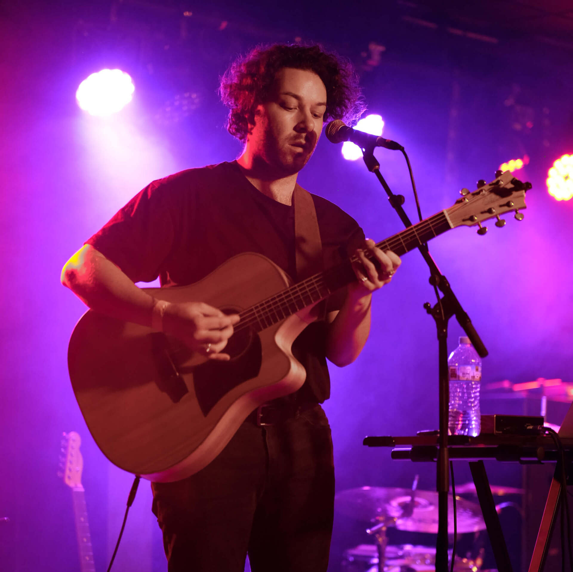 Man playing acoustic guitar on stage under purple and pink lights.