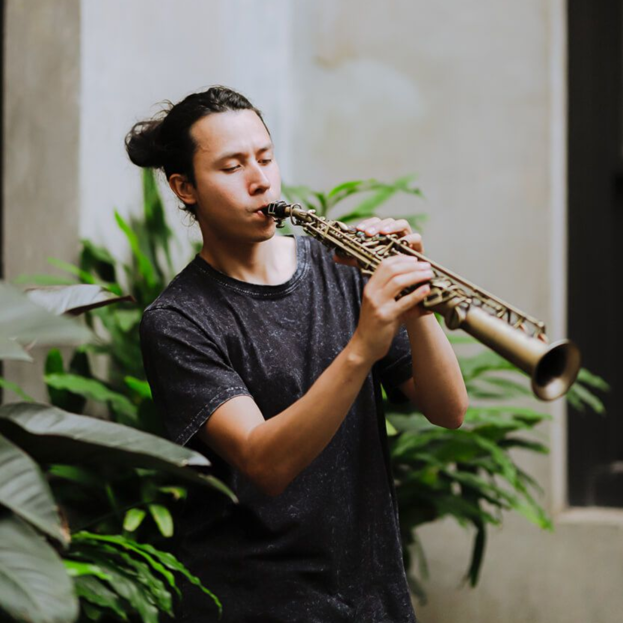 Ernesto playing a soprano saxophone, standing outdoors near greenery and a light-colored wall.