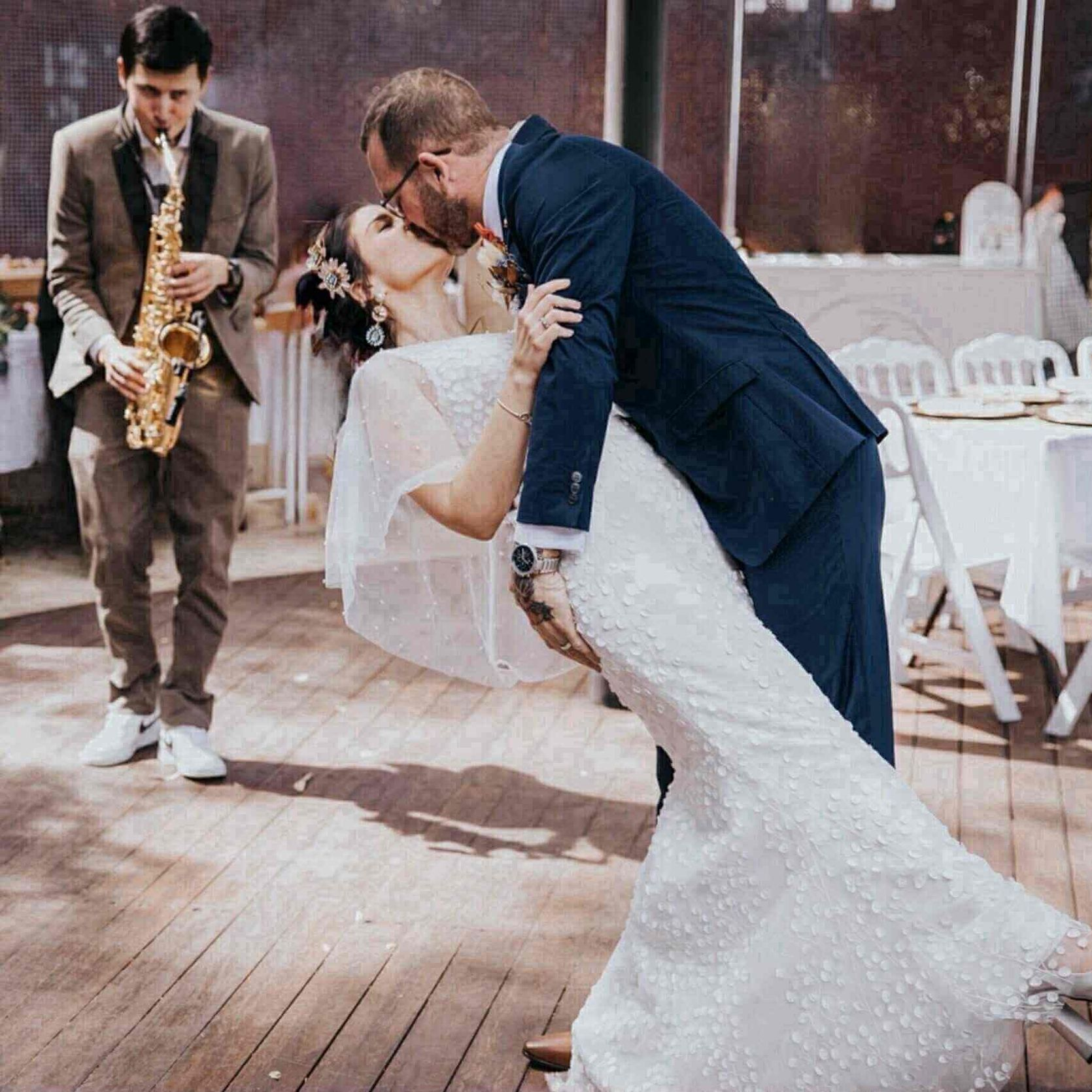 Bride and groom kissing, dancing on a wooden deck as a musician plays the saxophone at an outdoor wedding.