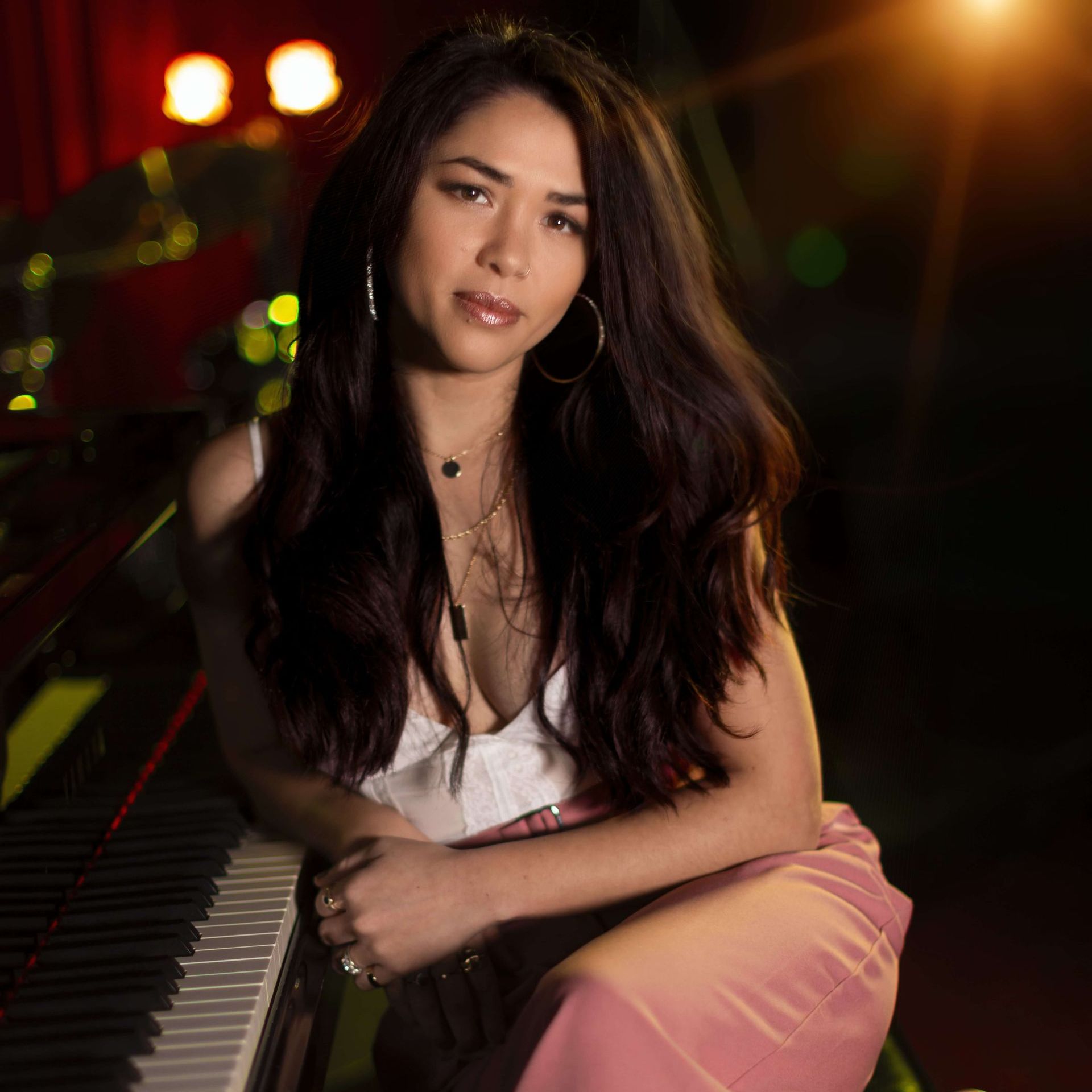 A woman with long, dark hair leans on a piano, looking at the camera. She wears a white top and pink pants; the background is dark with stage lights.