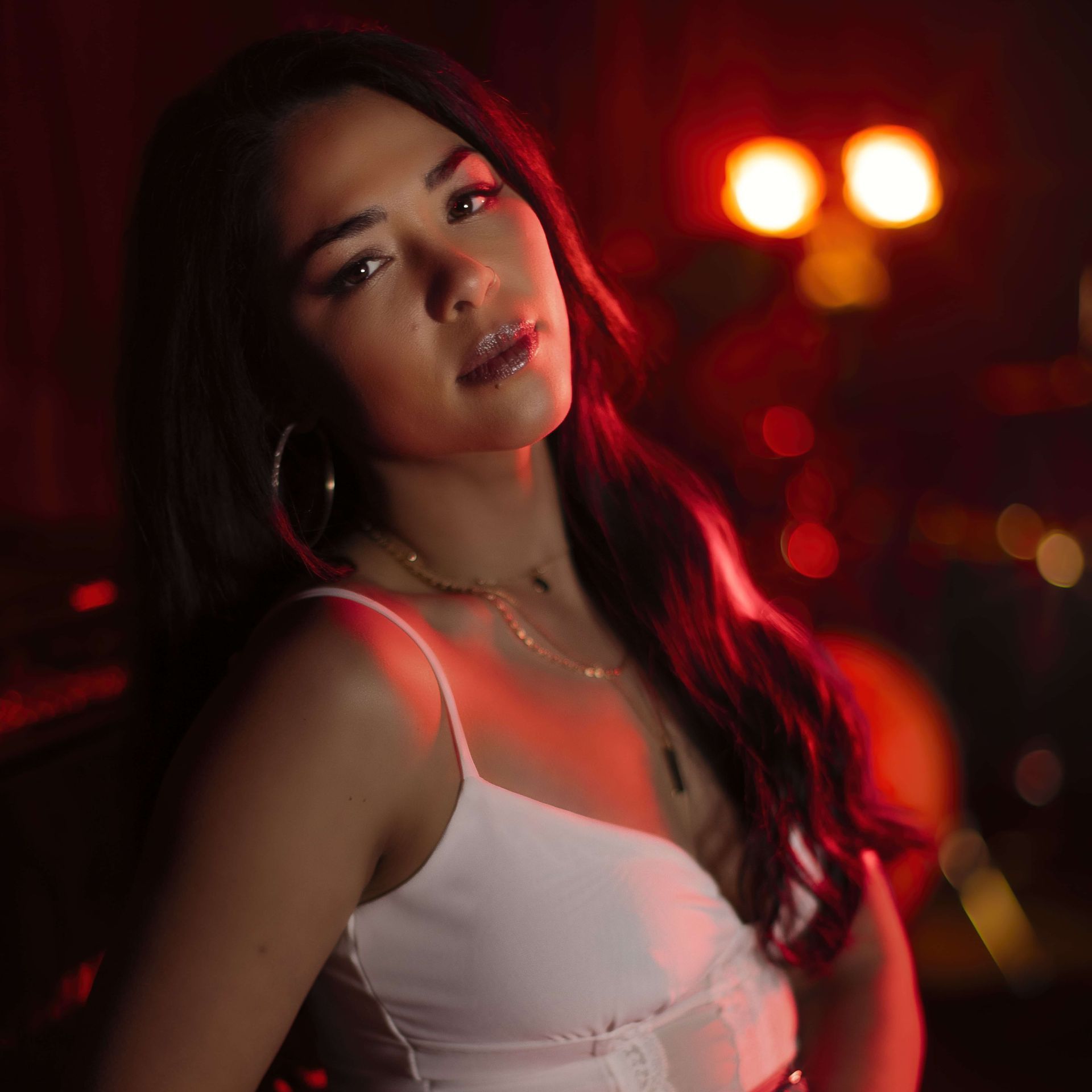 Young woman with long dark hair, wearing a white top, looking towards the camera in a dimly lit room with red and gold lighting.