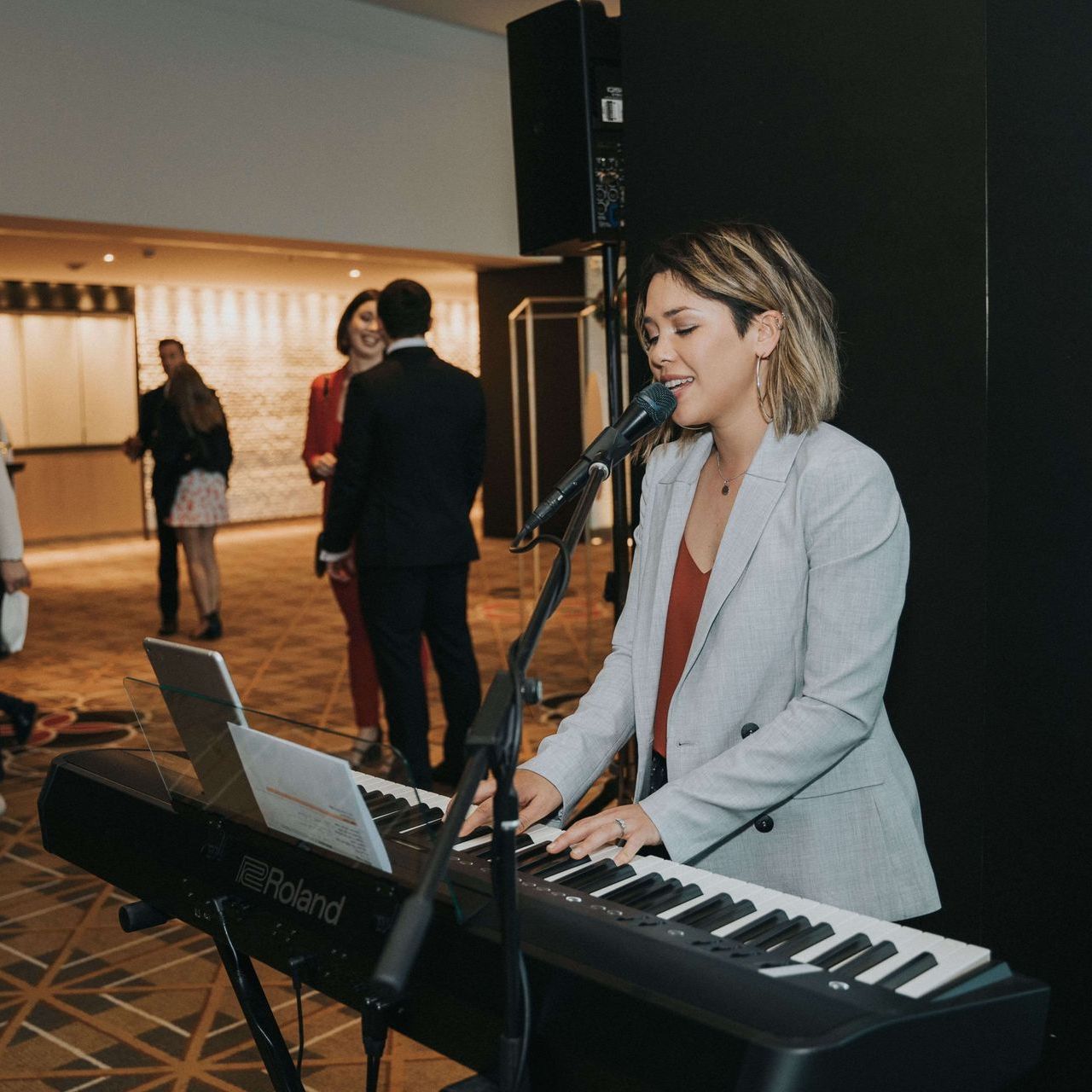 A woman sings and plays a Roland keyboard at an event. She wears a grey blazer and a red top. People mingle in the background.