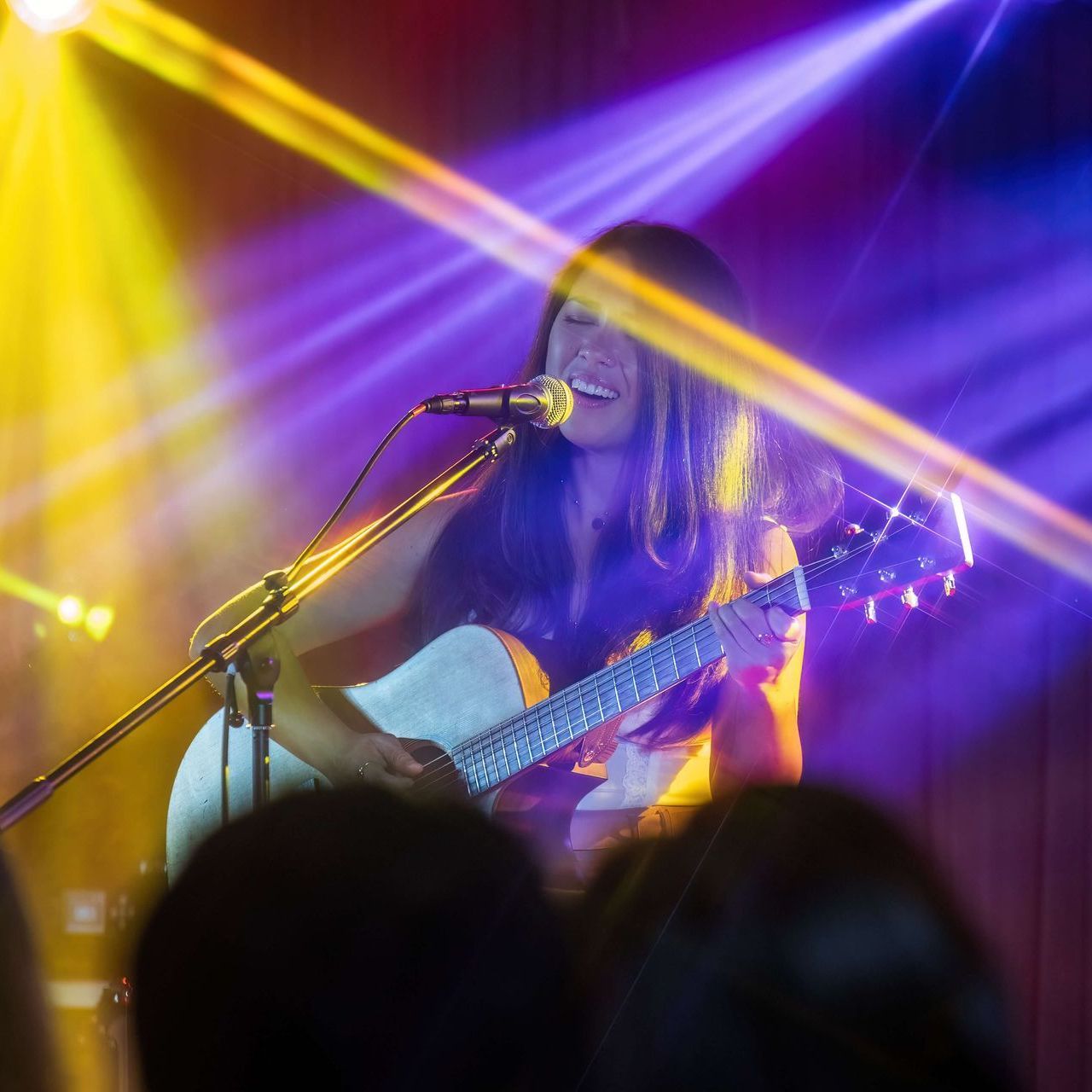 Woman singing and playing an acoustic guitar on stage, illuminated by colorful stage lights.