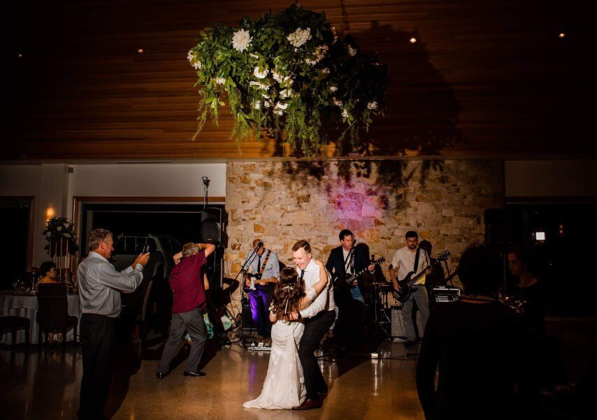A band performs on a stage with a brick wall backdrop; a singer stands between a saxophonist, bassist, and drummer.