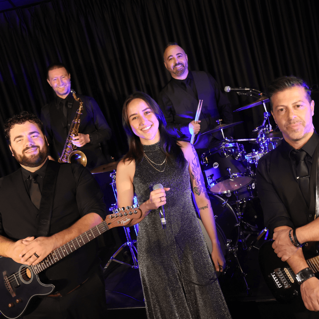 A band of five people in black outfits: a female singer, guitarist, saxophonist, drummer, and bass guitarist.