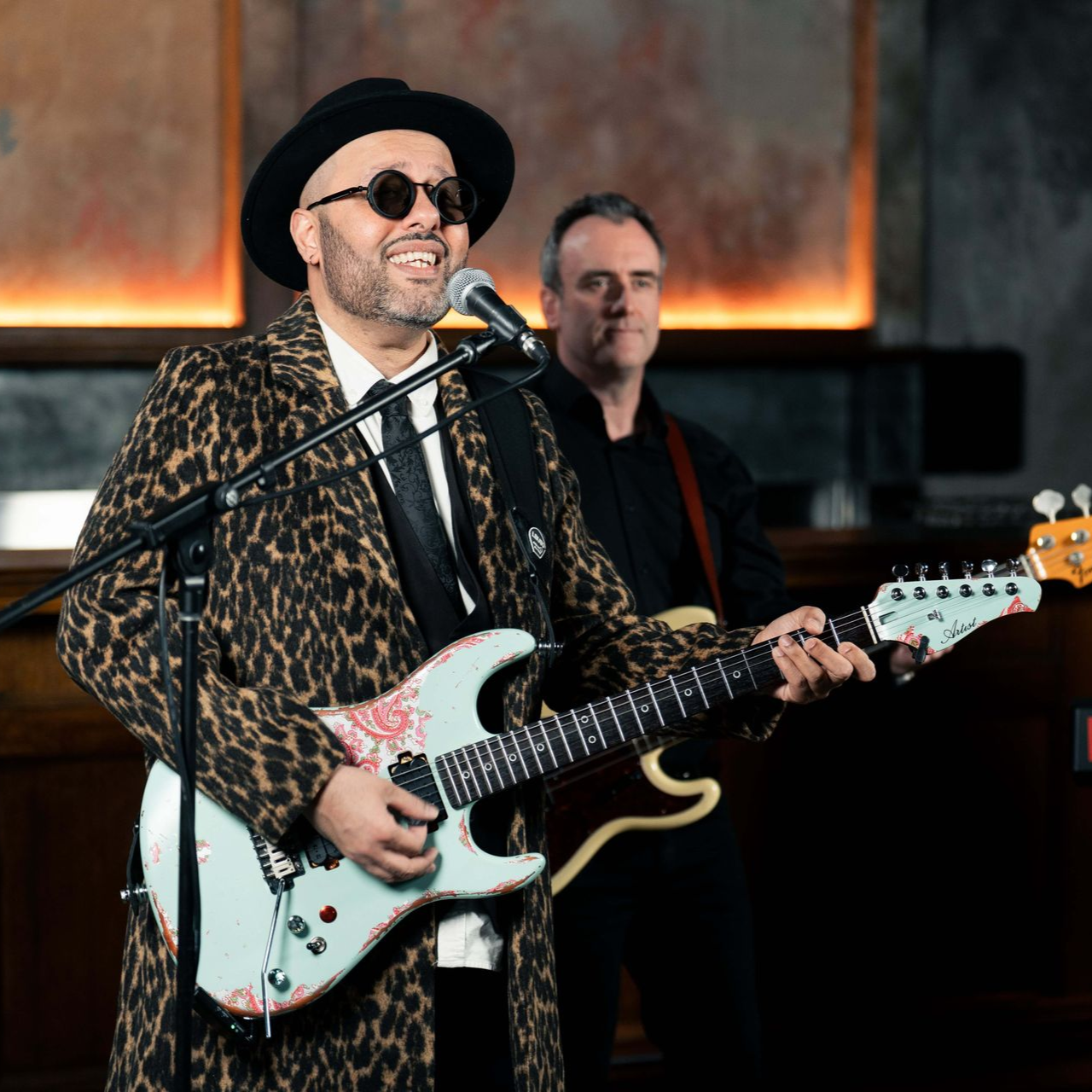 Man singing, playing guitar, in cheetah print coat; guitarist in background. Indoors.
