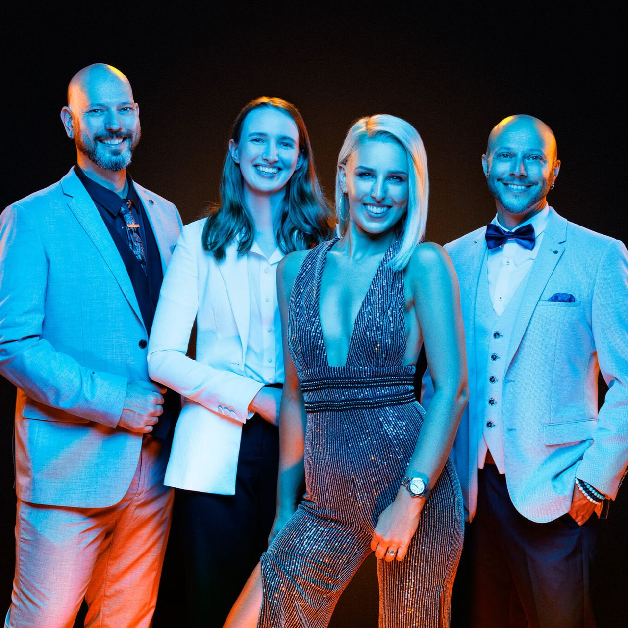Four people in formal attire posing against a dark background, lit with blue and orange lights.