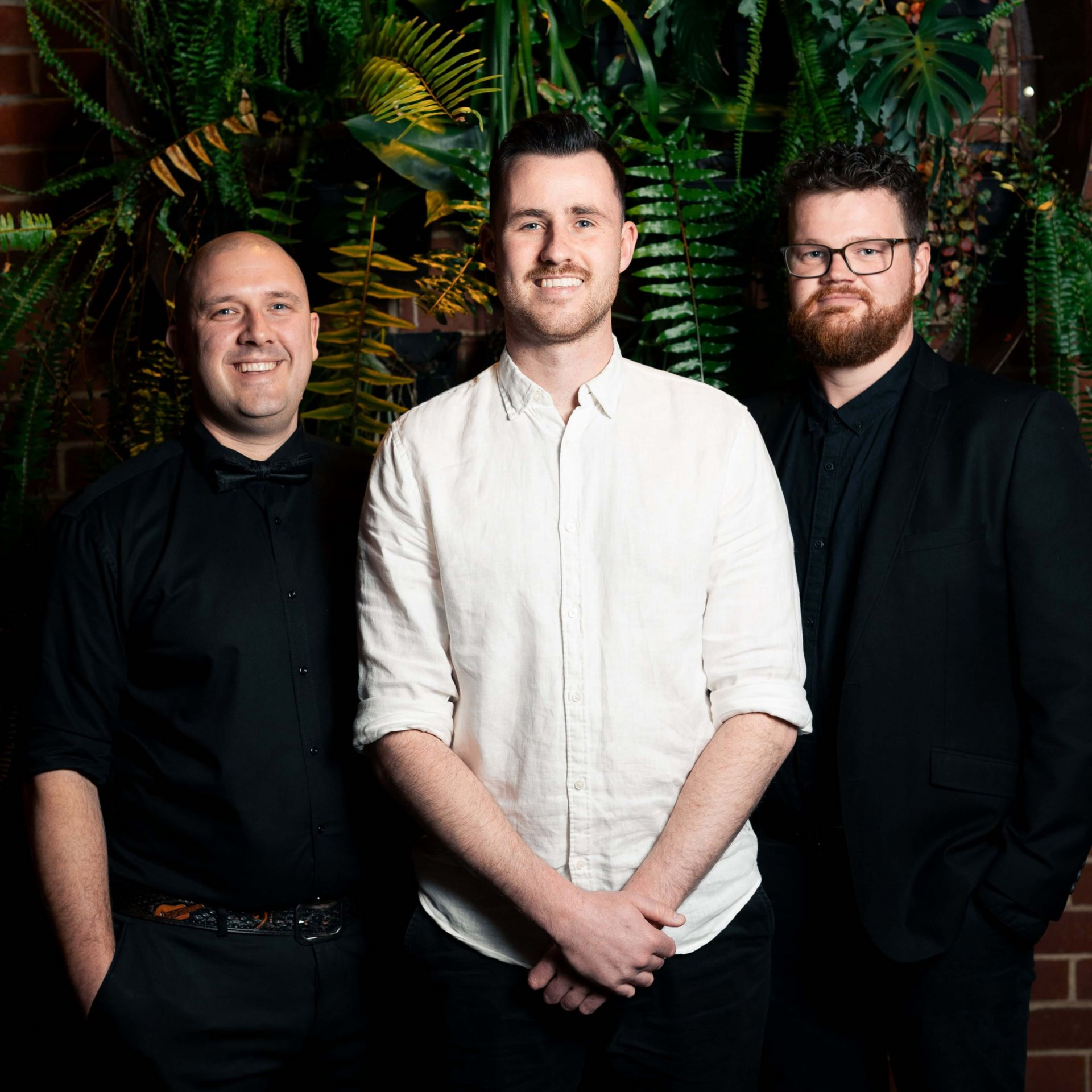 Three men in a room with plants, smiling at the camera. One in a white shirt, two in black.