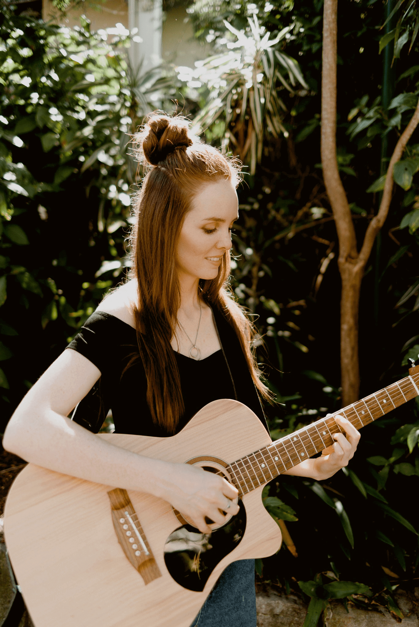 A woman is playing an acoustic guitar in front of a tree.