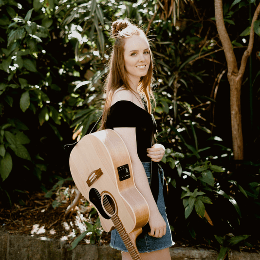 A woman is holding an acoustic guitar in front of trees.