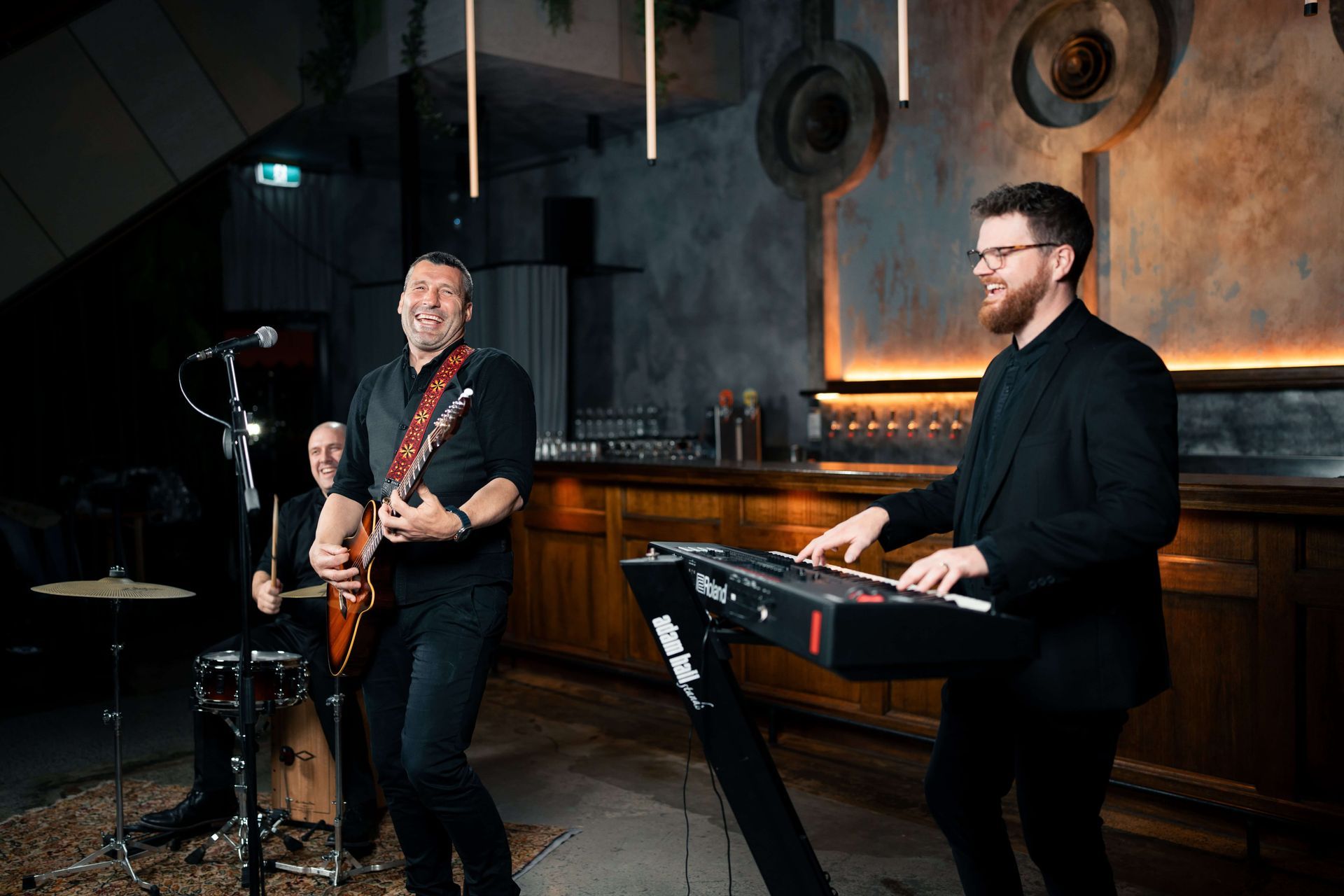 A band performs on a stage with a brick wall backdrop; a singer stands between a saxophonist, bassist, and drummer.