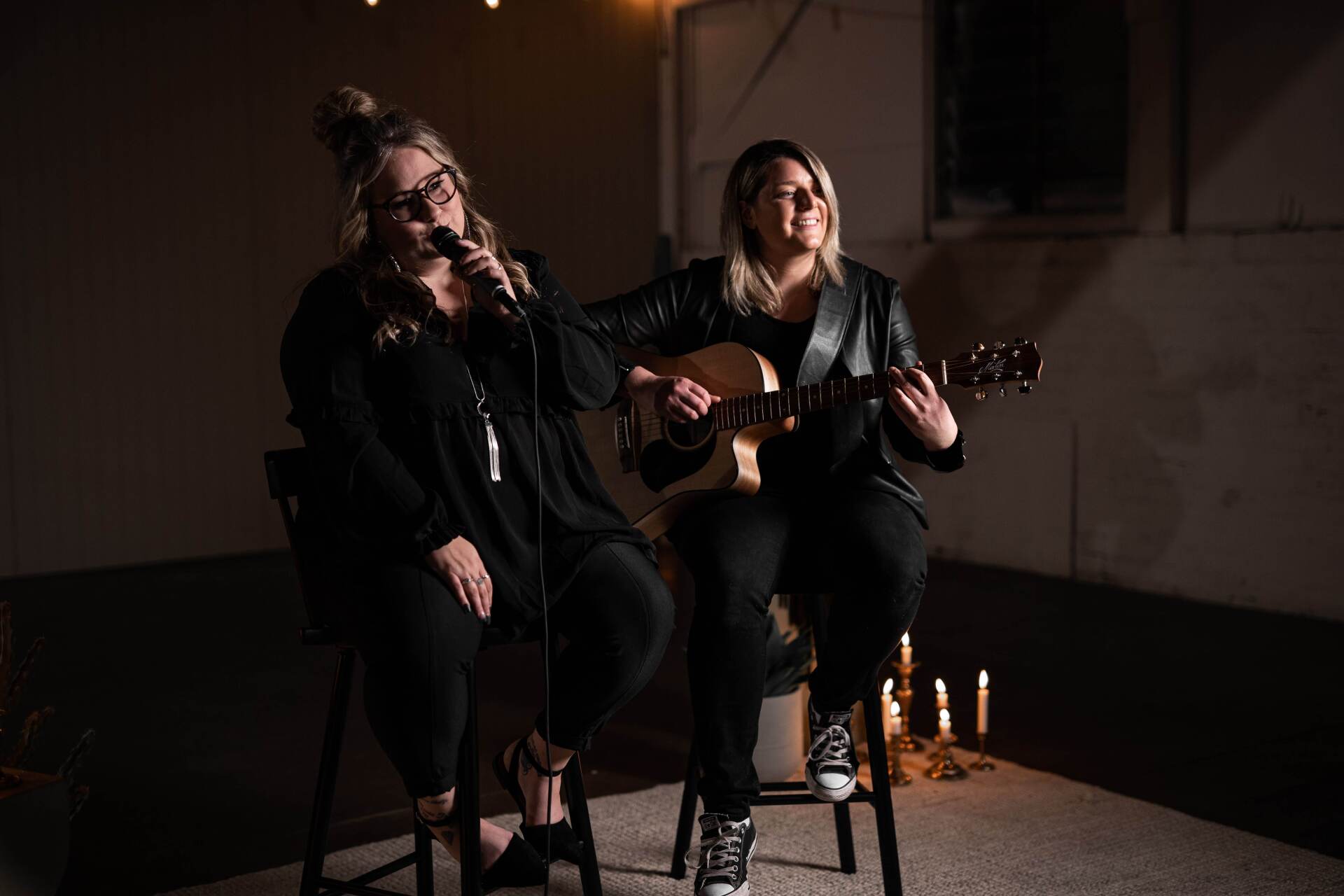 Two women performing music. One sings, the other plays guitar. Dark setting, lit by candles.